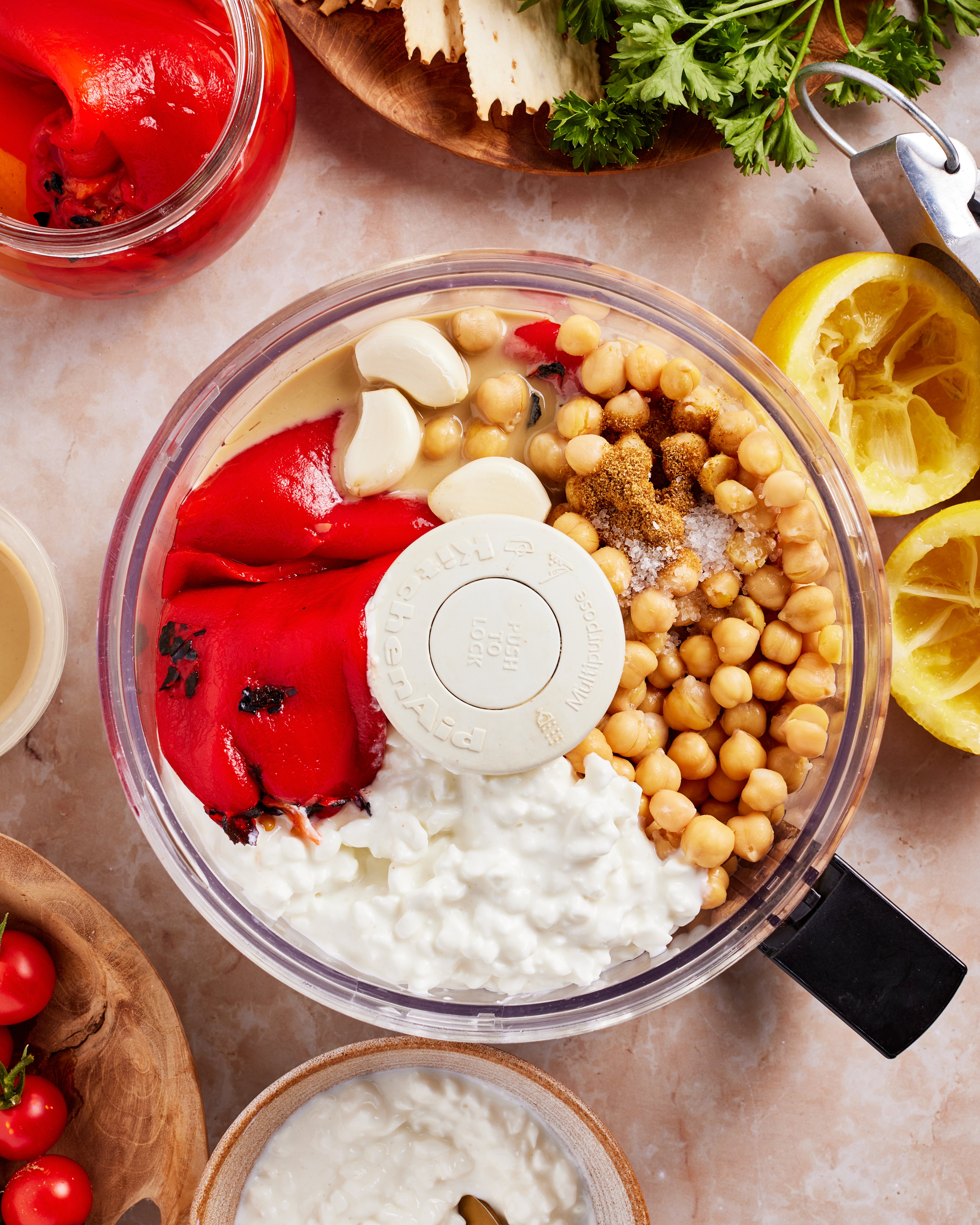 A food processor filled with chickpeas, roasted red peppers, garlic cloves, cottage cheese, tahini, and spices sits on a countertop surrounded by lemon halves, parsley, tomatoes, and other ingredients.
