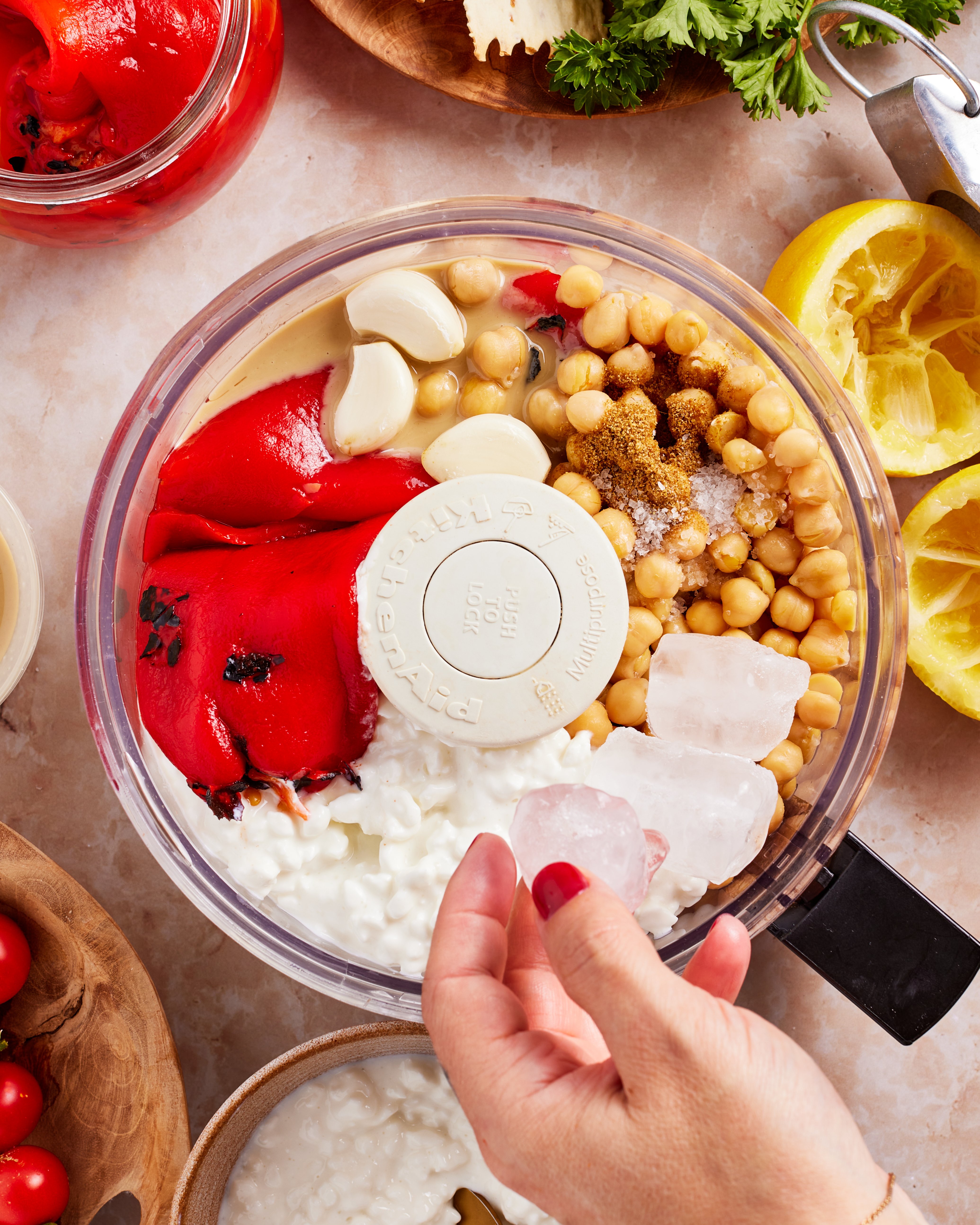 A hand adds ice cubes to a food processor filled with chickpeas, roasted red peppers, cottage cheese, garlic, tahini, brown sugar, and seasonings on a kitchen counter with ingredients nearby.
