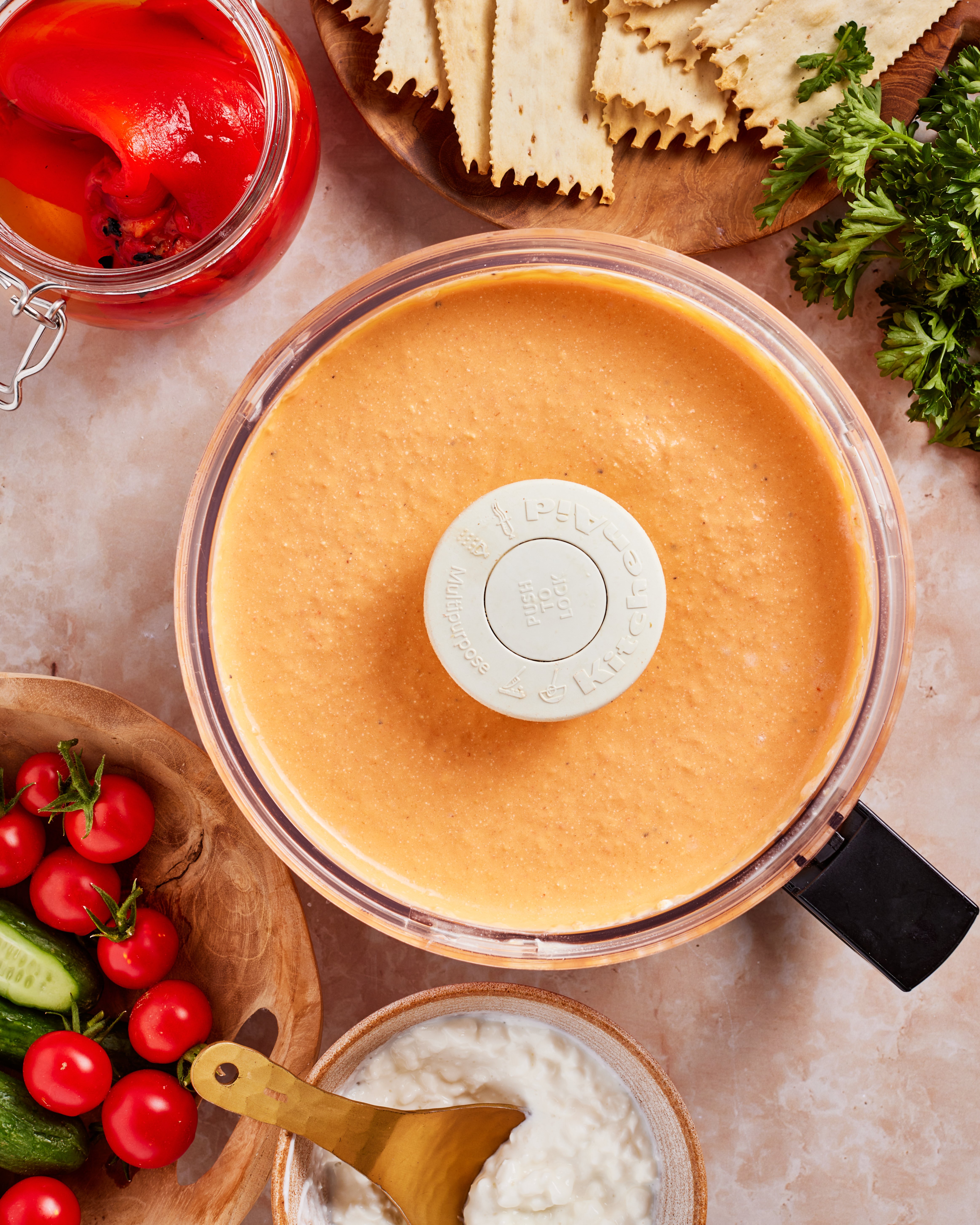 A food processor filled with creamy orange dip sits on a countertop surrounded by crackers, parsley, jarred red peppers, cherry tomatoes, cucumbers, and a bowl of cottage cheese with a gold spoon.