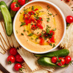 A bowl of creamy dip garnished with chopped herbs, diced red peppers, and a drizzle of olive oil, surrounded by sliced cucumbers, cherry tomatoes, and rectangular crackers on a plate.
