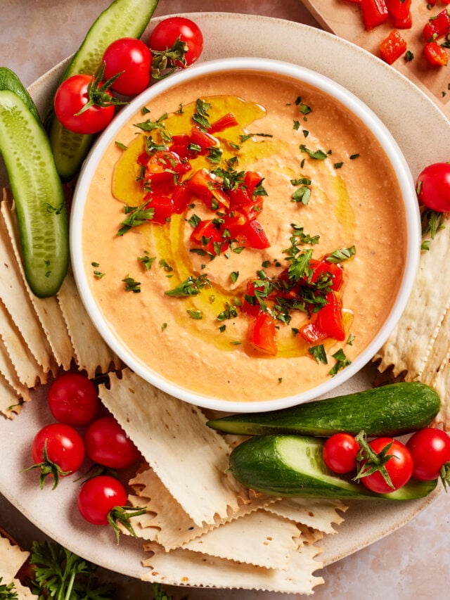 A bowl of creamy dip garnished with chopped herbs, diced red peppers, and a drizzle of olive oil, surrounded by sliced cucumbers, cherry tomatoes, and rectangular crackers on a plate.