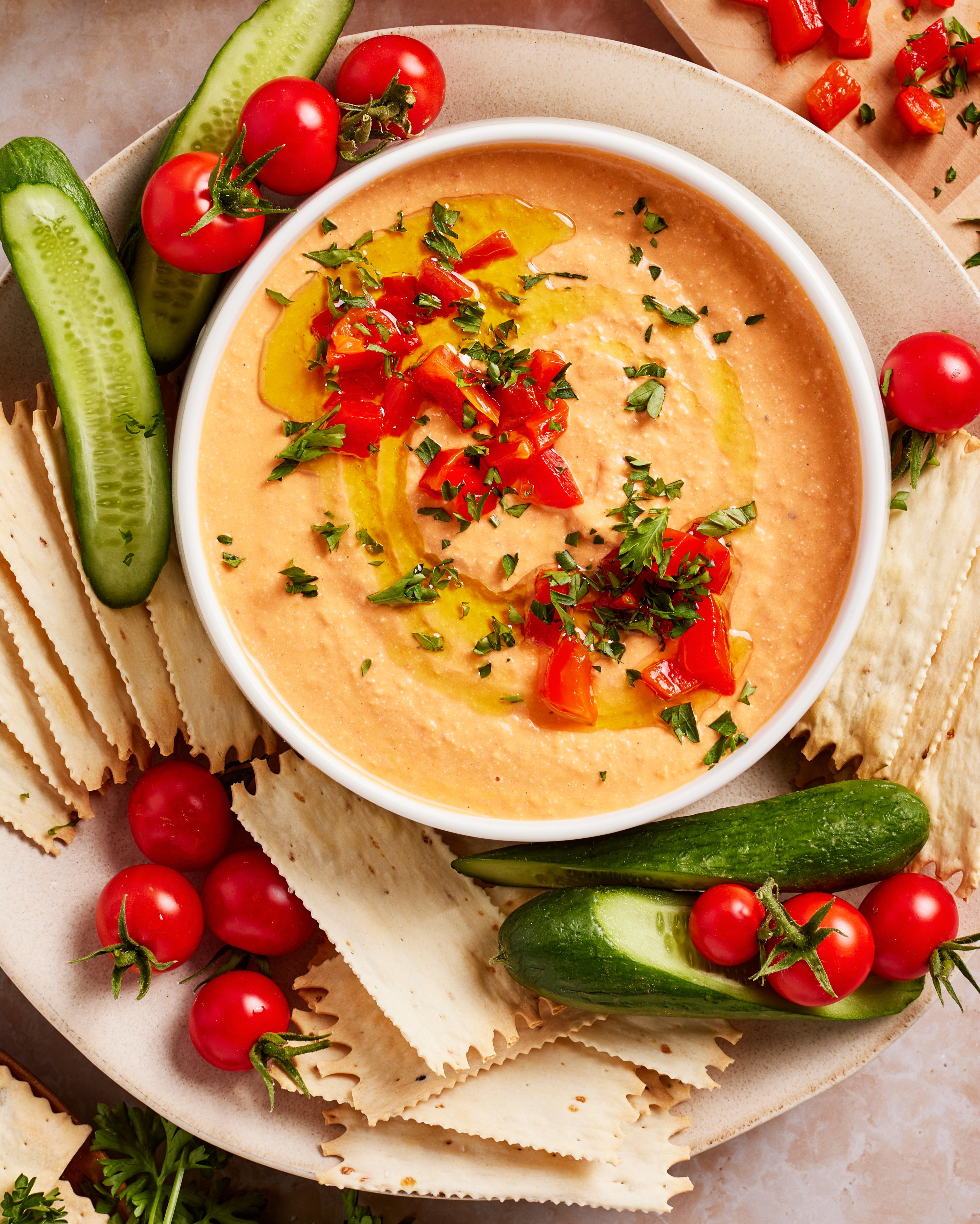 A bowl of creamy dip garnished with chopped herbs, diced red peppers, and a drizzle of olive oil, surrounded by sliced cucumbers, cherry tomatoes, and rectangular crackers on a plate.