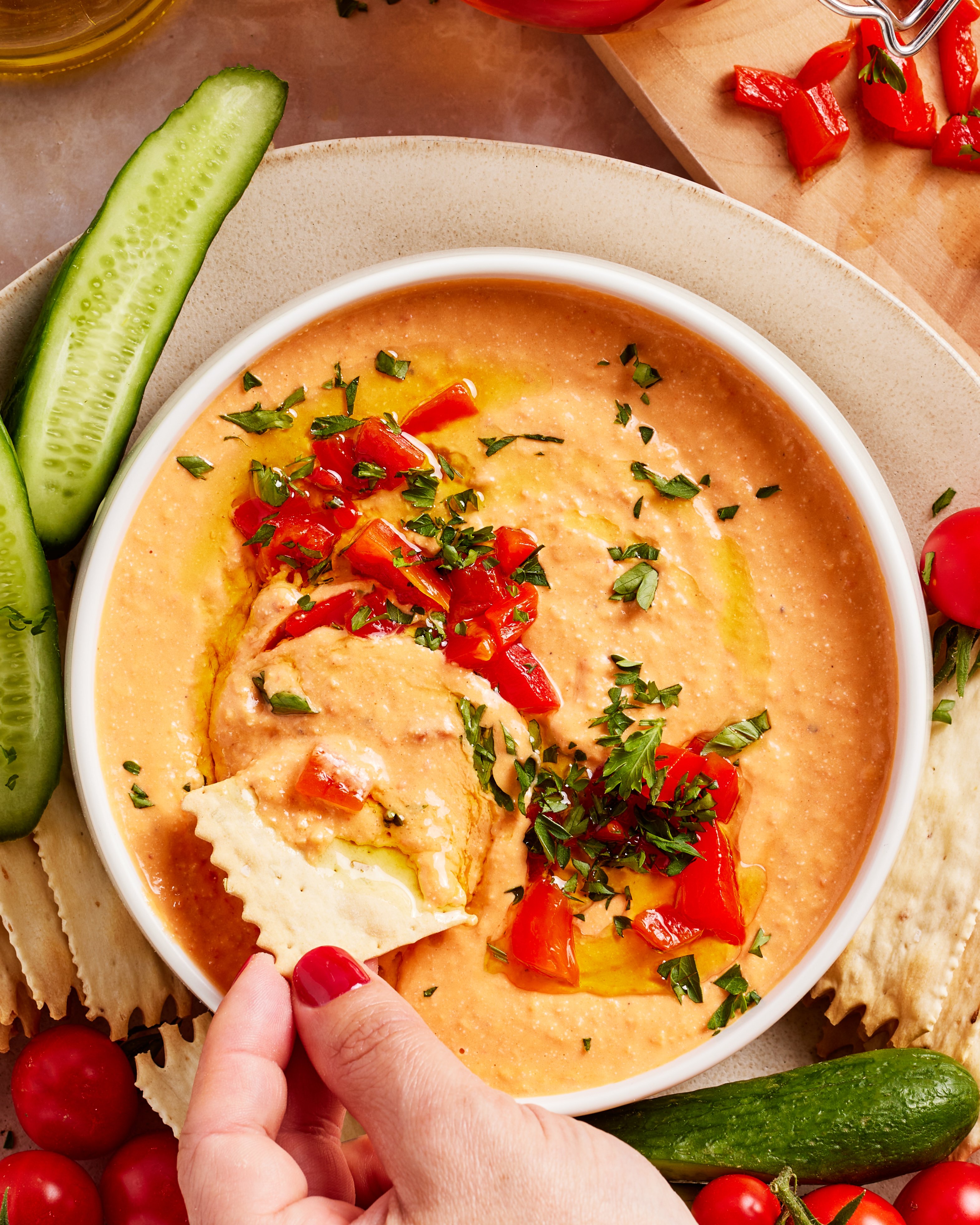 A hand dips a cracker into a bowl of creamy red pepper hummus, garnished with chopped herbs and red peppers. Sliced cucumbers, cherry tomatoes, and more crackers surround the bowl on a plate.