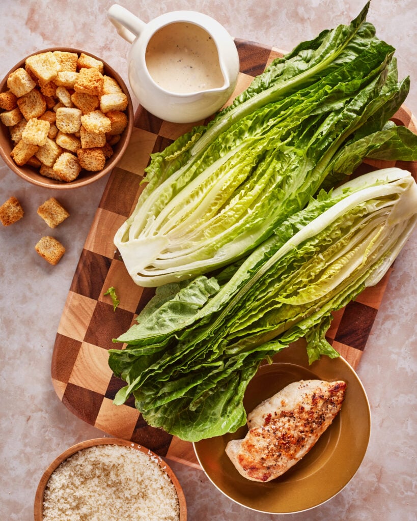 A top view of Caesar salad ingredients: halved romaine lettuce, a bowl of croutons, a small bowl of grated cheese, grilled chicken breast on a plate, and a jug of Caesar dressing on a checkered cutting board.