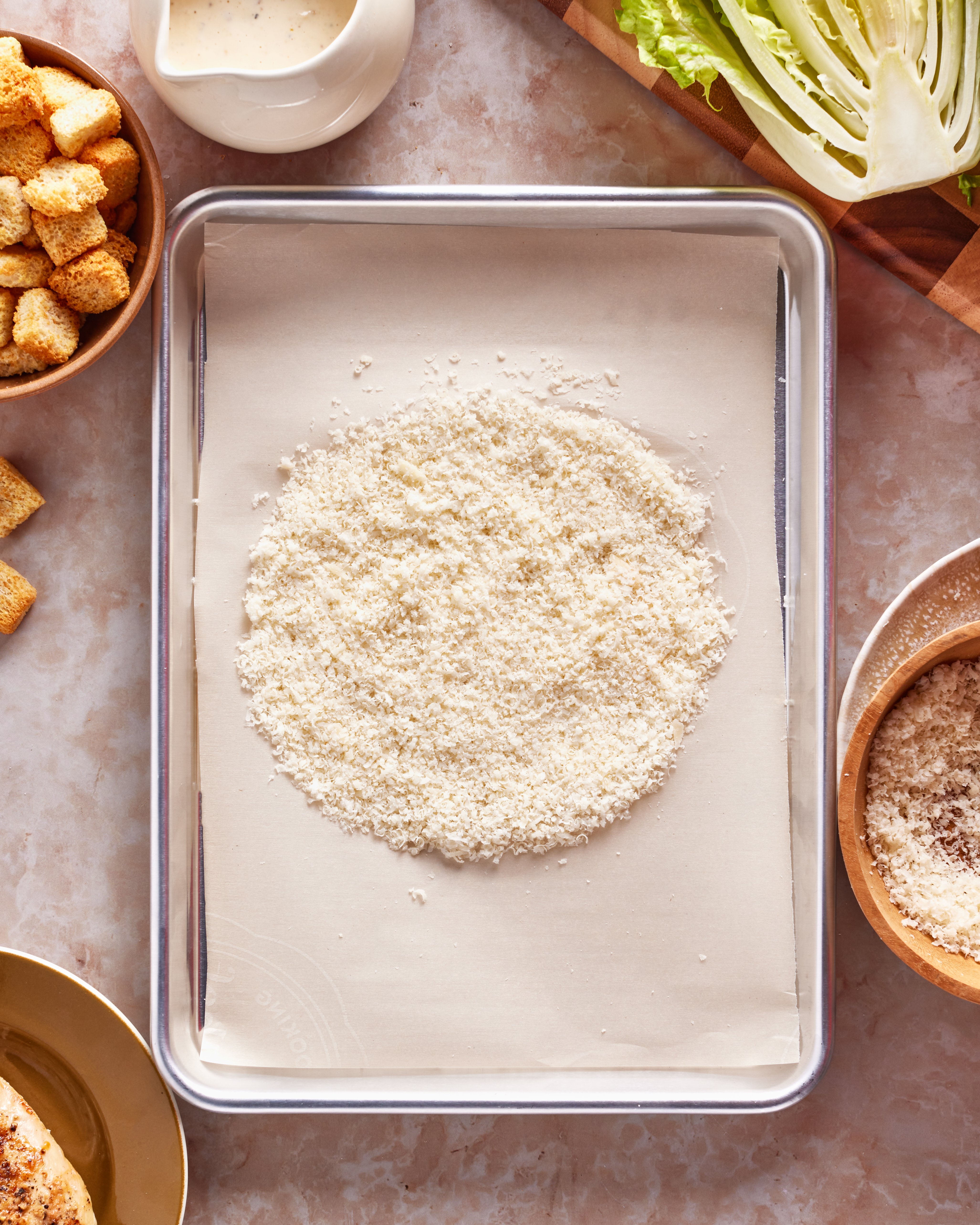 A parchment-lined baking sheet with a mound of breadcrumbs in the center, surrounded by bowls of croutons, grated cheese, lettuce wedges, and a container of dressing on a light countertop.