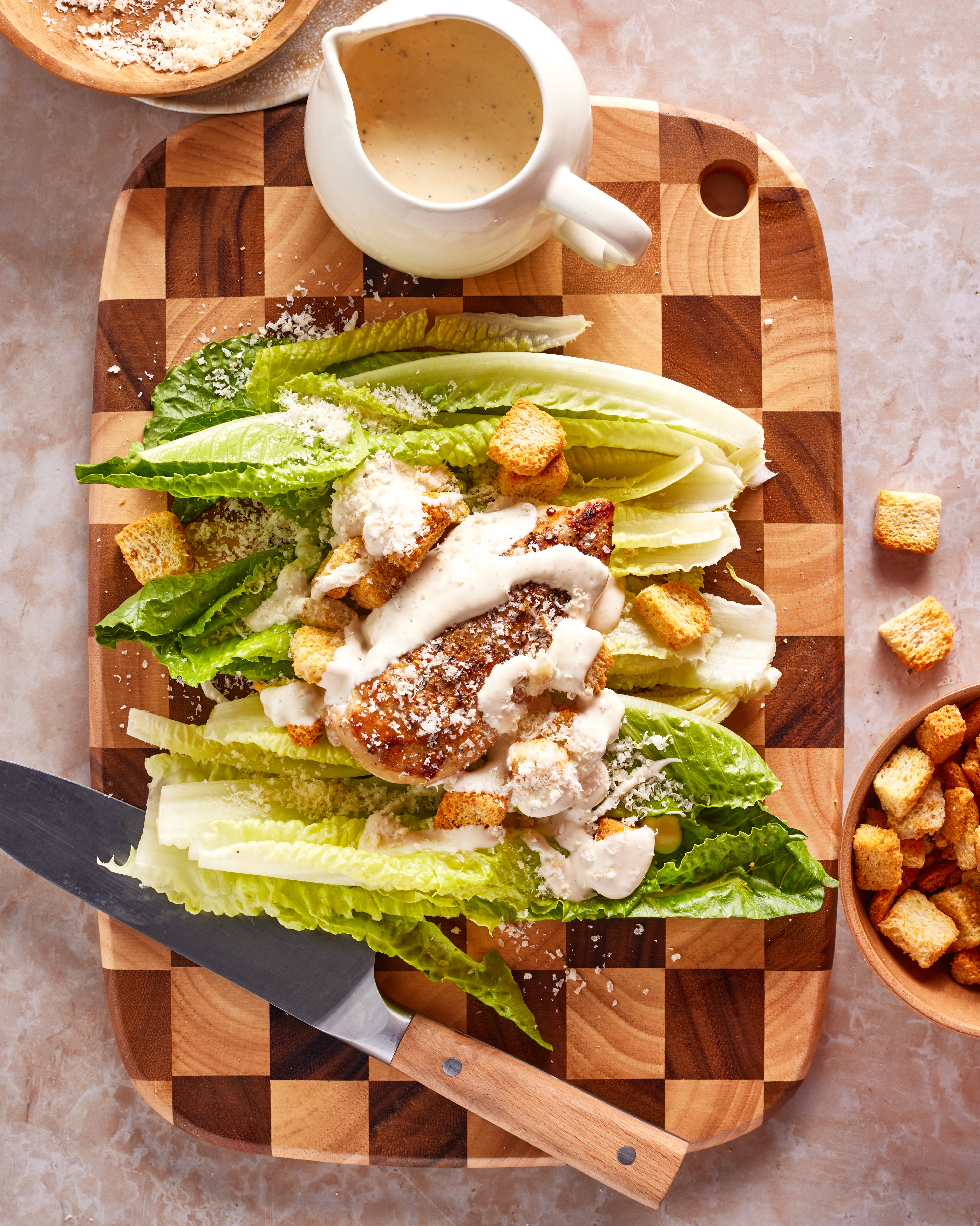 A Caesar salad with grilled chicken, romaine lettuce, croutons, and Caesar dressing is arranged on a wooden cutting board. A knife, a small bowl of croutons, and a pitcher of dressing are nearby.