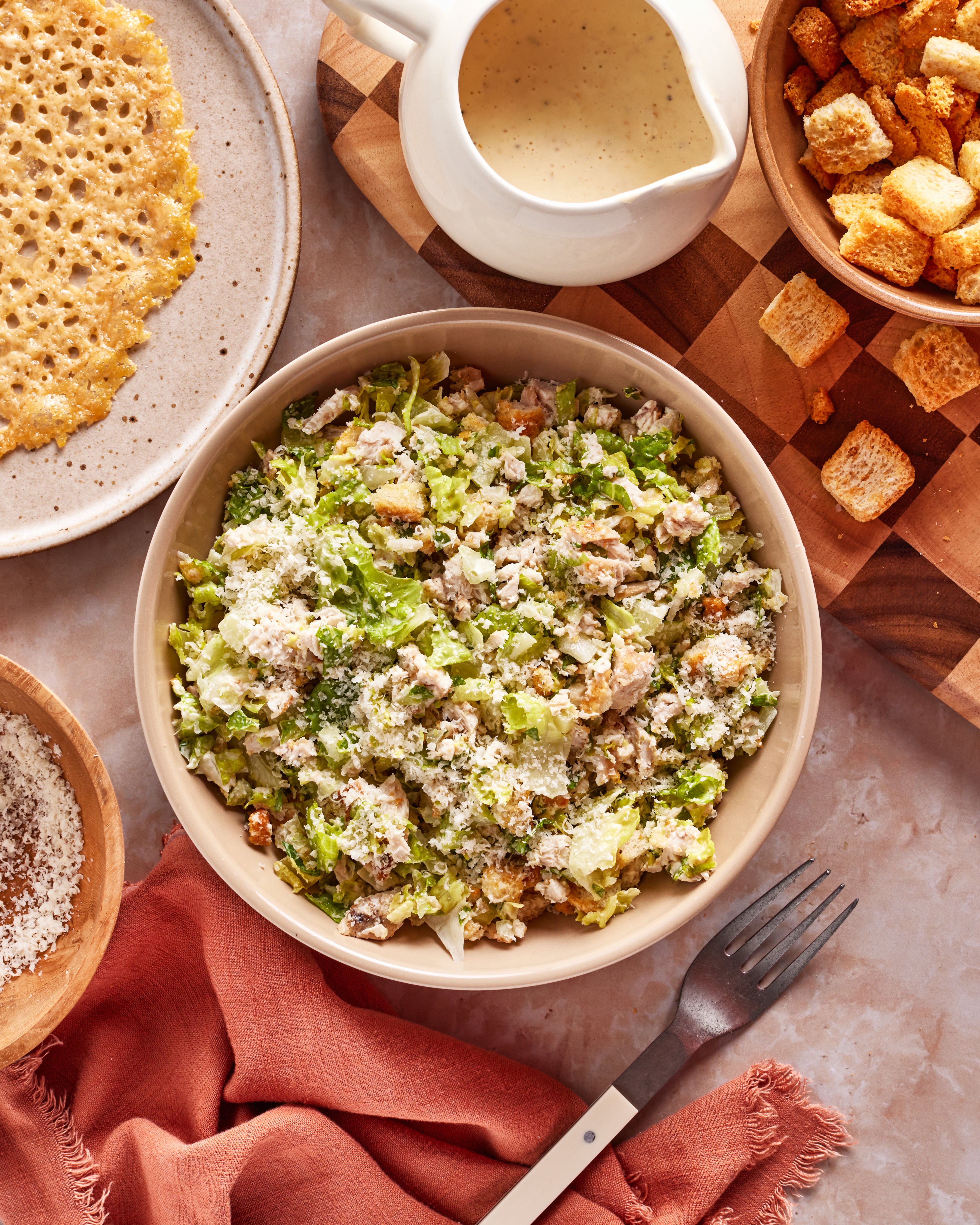 A bowl of Caesar salad topped with grated cheese sits on a table, surrounded by a plate of cheese crisps, a jug of dressing, a bowl of croutons, grated cheese, a fork, and a rust-colored napkin.