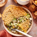 A bowl of Caesar salad with grilled chicken is topped with a large, lacy Parmesan crisp. A fork rests inside the bowl, and Caesar dressing, croutons, and grated cheese are placed nearby on the table.