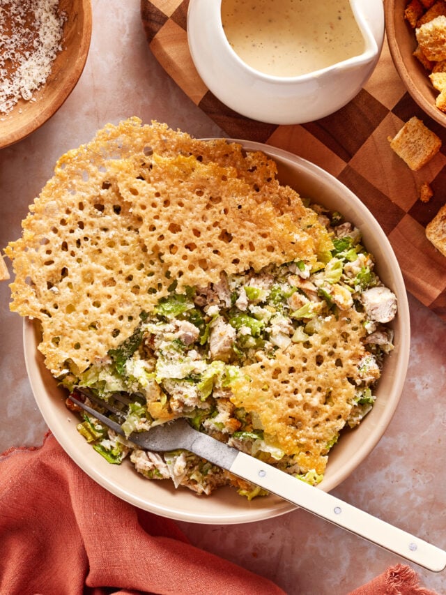 A bowl of Caesar salad with grilled chicken is topped with a large, lacy Parmesan crisp. A fork rests inside the bowl, and Caesar dressing, croutons, and grated cheese are placed nearby on the table.