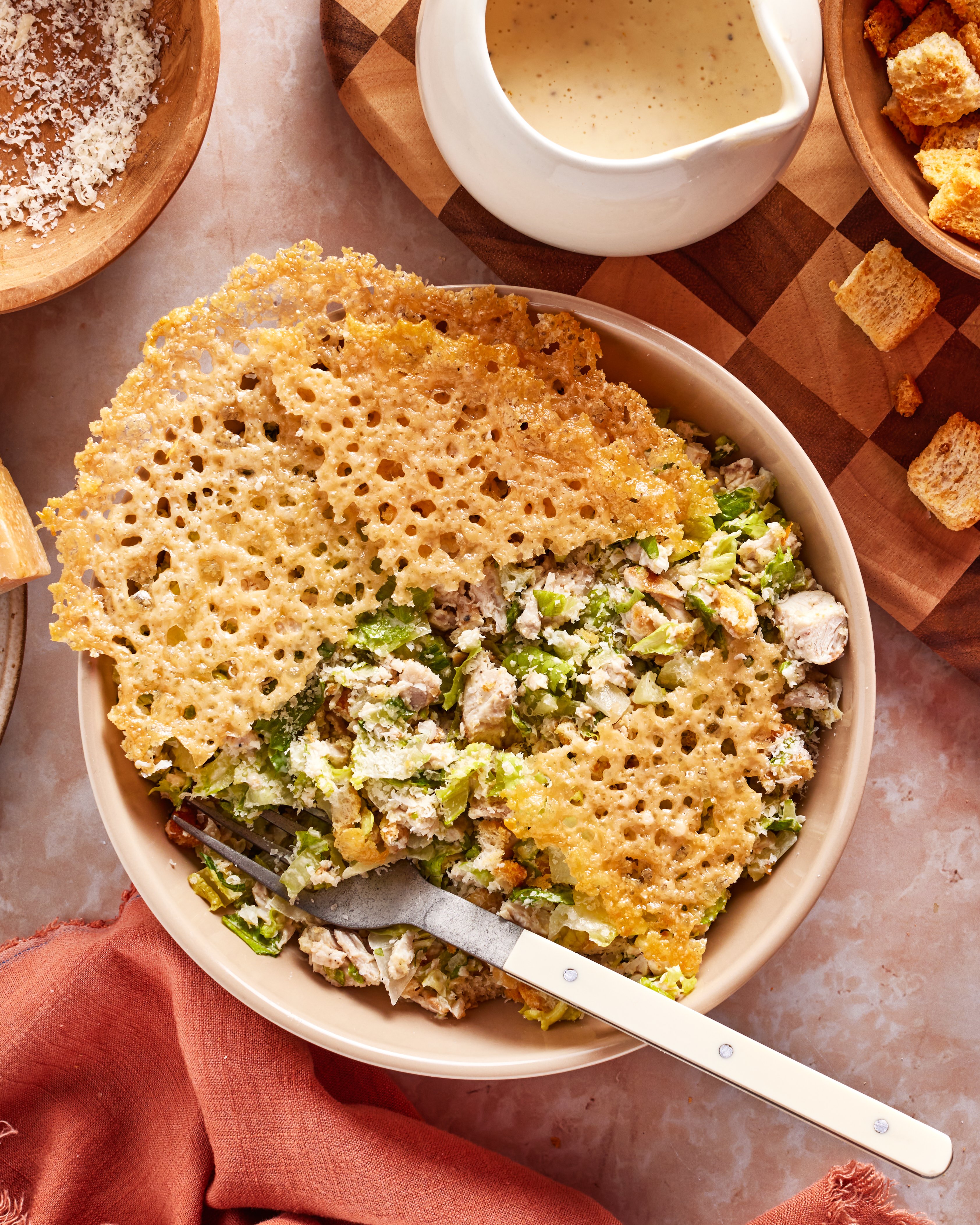 A bowl of Caesar salad with grilled chicken is topped with a large, lacy Parmesan crisp. A fork rests inside the bowl, and Caesar dressing, croutons, and grated cheese are placed nearby on the table.