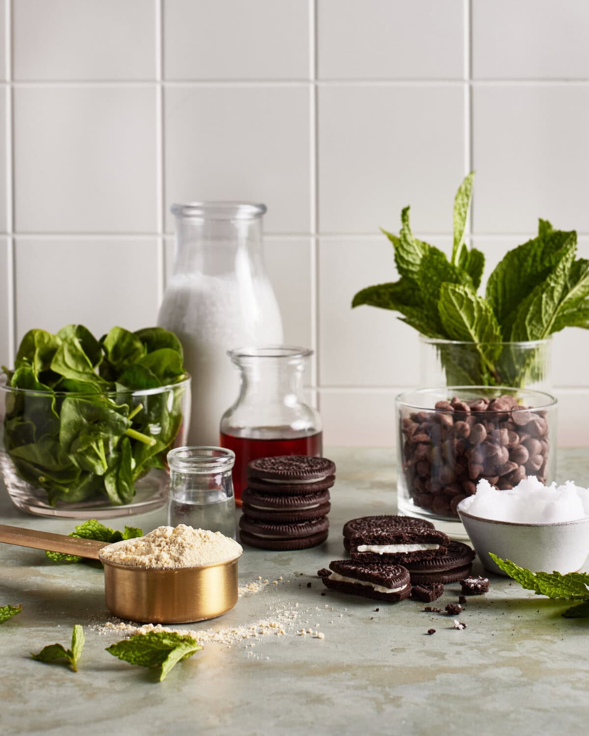 A countertop displays spinach leaves, a jug of milk, a glass of red liquid, cocoa beans, fresh mint, a small jar of powder, chocolate sandwich cookies, a measuring cup with flour, and a bowl of coconut oil.