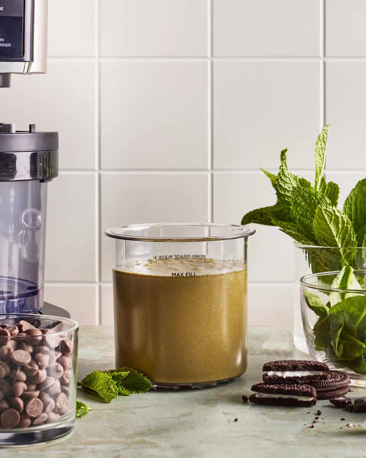 A glass blender container filled with a greenish smoothie sits on a counter next to a small bowl of chocolate chips, a few chocolate sandwich cookies, and fresh mint leaves, with a blender and white tile background visible.