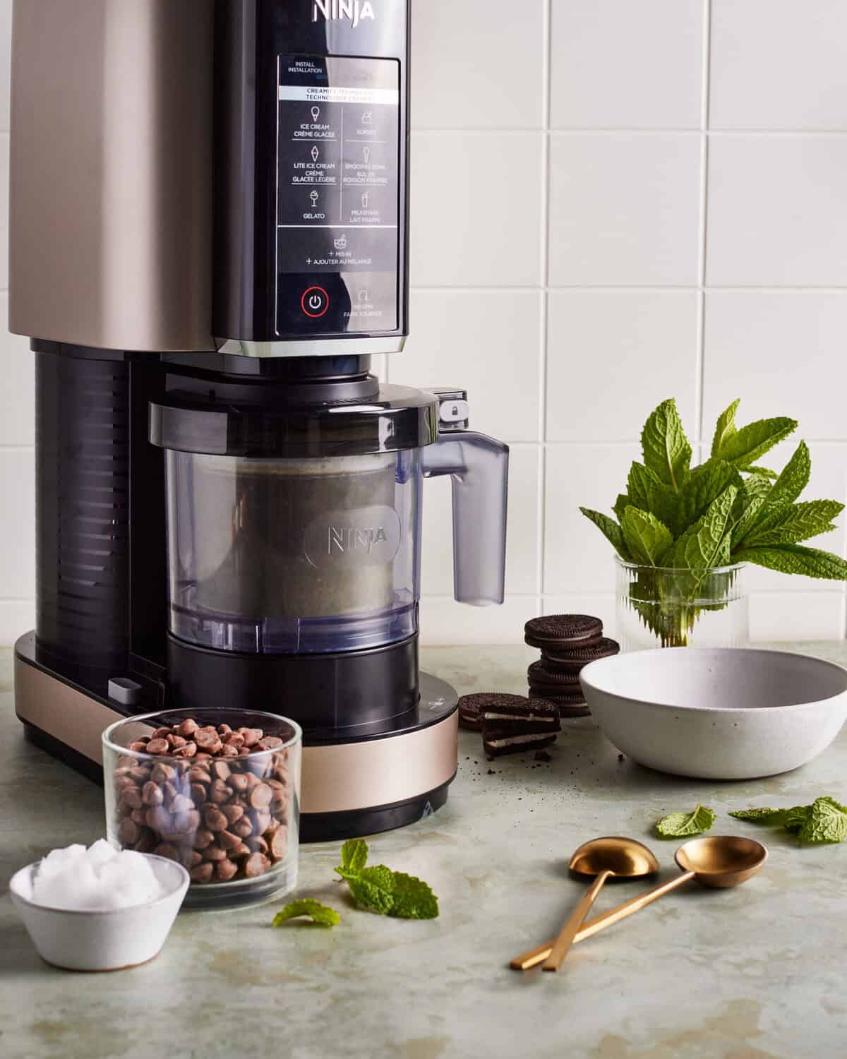 A Ninja ice cream maker sits on a kitchen counter with a glass of chocolate chips, a bowl of white powder, cookies, fresh mint leaves, a gold spoon and fork, and a mint plant in the background.