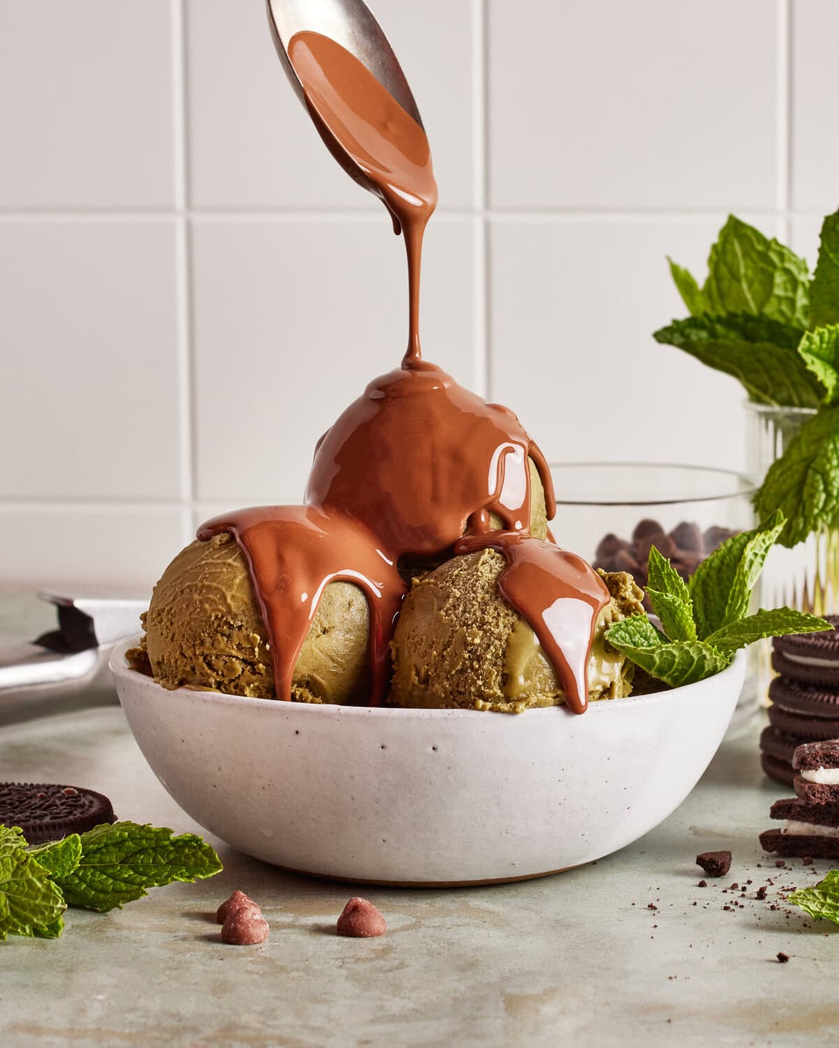 A spoon drizzles chocolate sauce over three scoops of ice cream in a white bowl. Mint sprigs, chocolate chips, and sandwich cookies are arranged around the bowl on a light surface.
