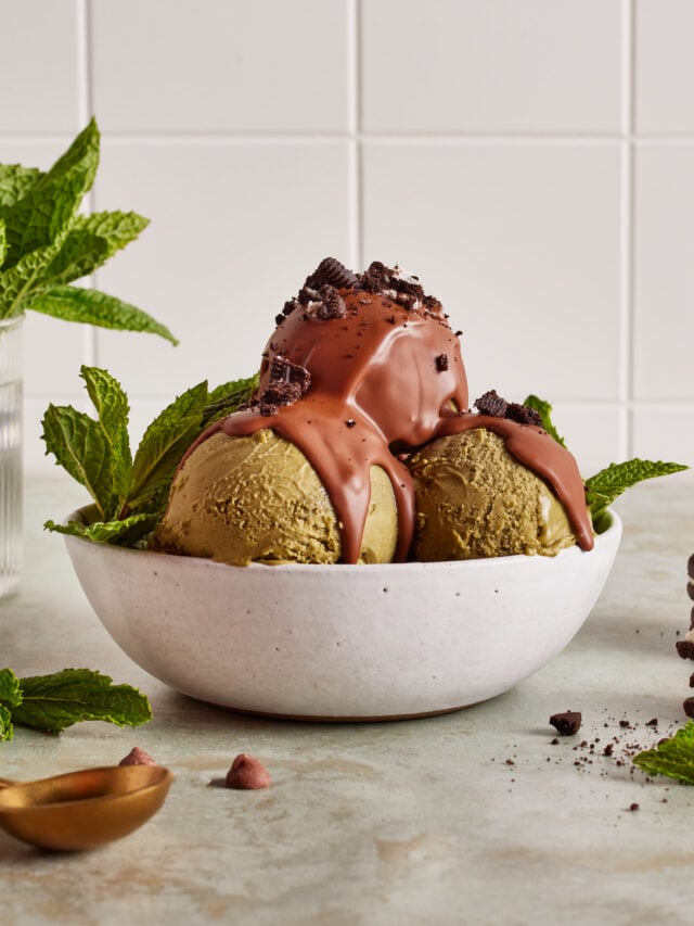 Two scoops of green ice cream in a white bowl, topped with chocolate sauce and cookie crumbles, garnished with mint leaves. A vase with mint and a stack of cookies are in the background.