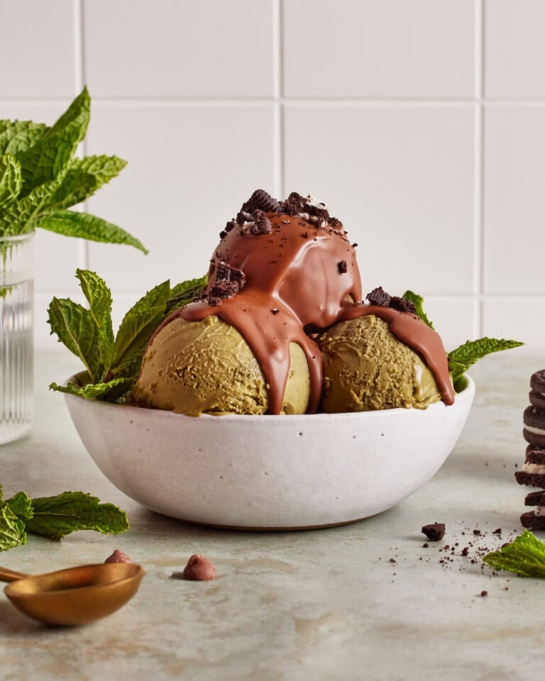 Two scoops of green ice cream in a white bowl, topped with chocolate sauce and cookie crumbles, garnished with mint leaves. A vase with mint and a stack of cookies are in the background.