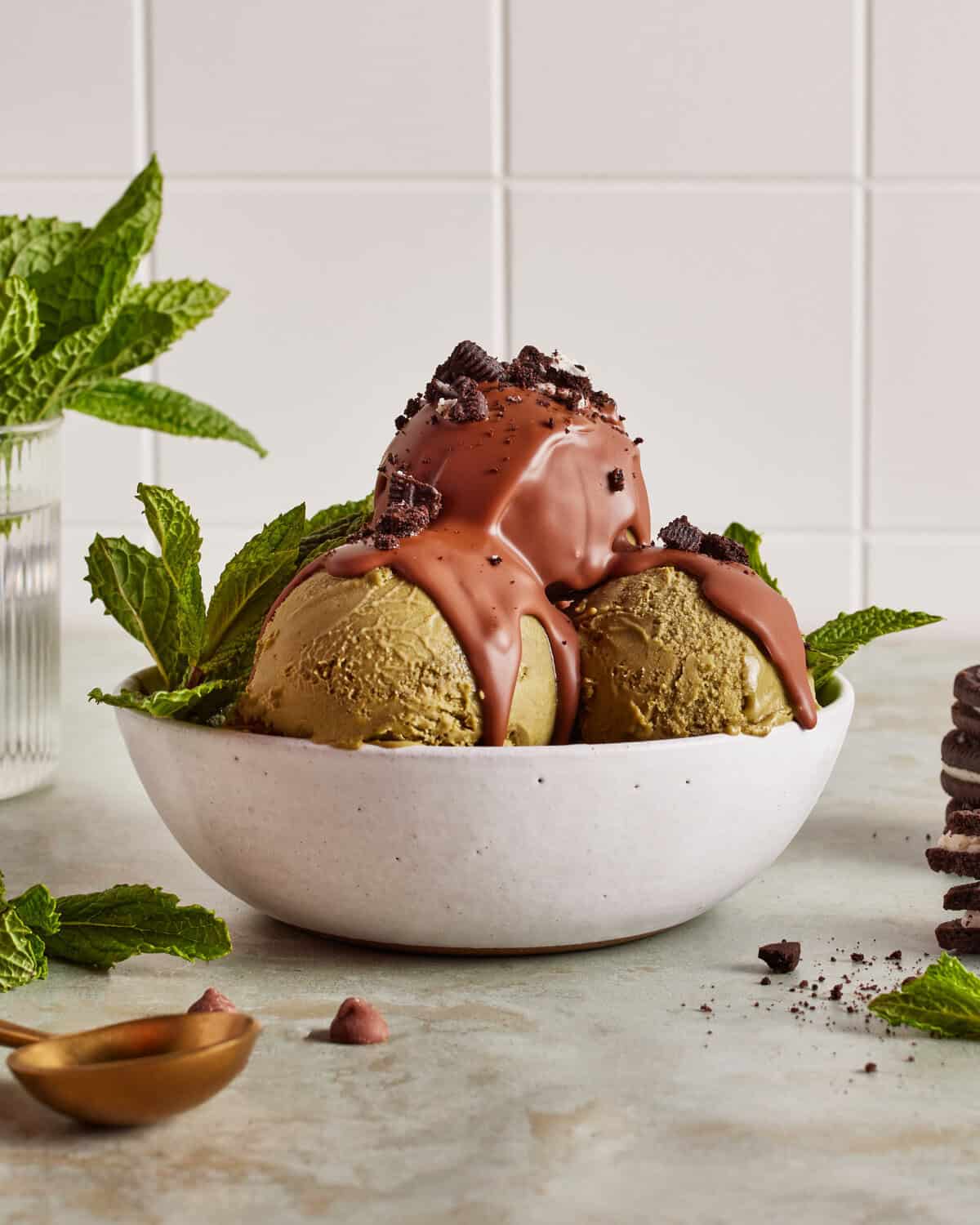 Two scoops of green ice cream in a white bowl, topped with chocolate sauce and cookie crumbles, garnished with mint leaves. A vase with mint and a stack of cookies are in the background.