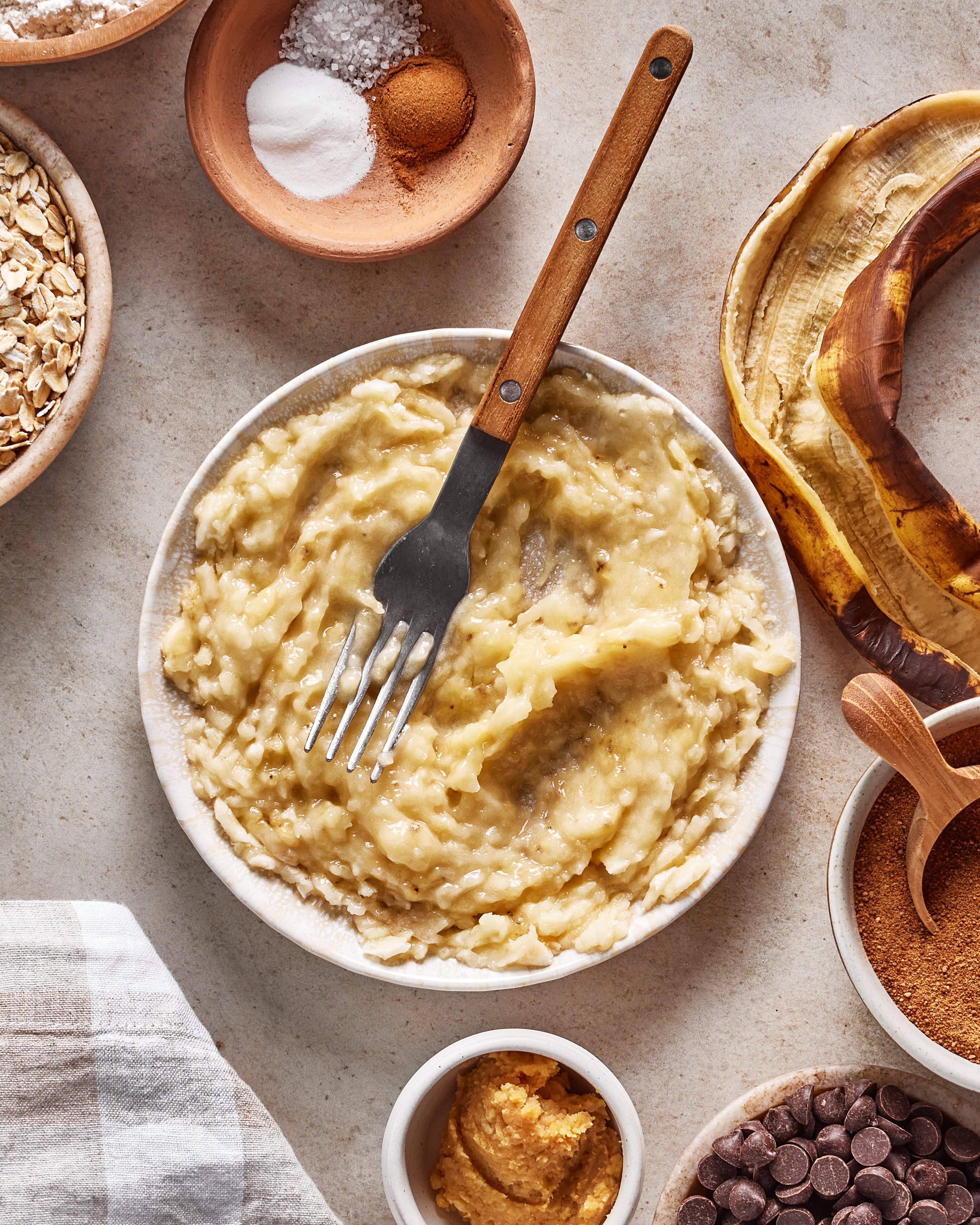 A bowl of mashed bananas with a fork, surrounded by baking ingredients: rolled oats, salt, cinnamon, sugar, peanut butter, chocolate chips, and a banana peel on a light-colored surface.