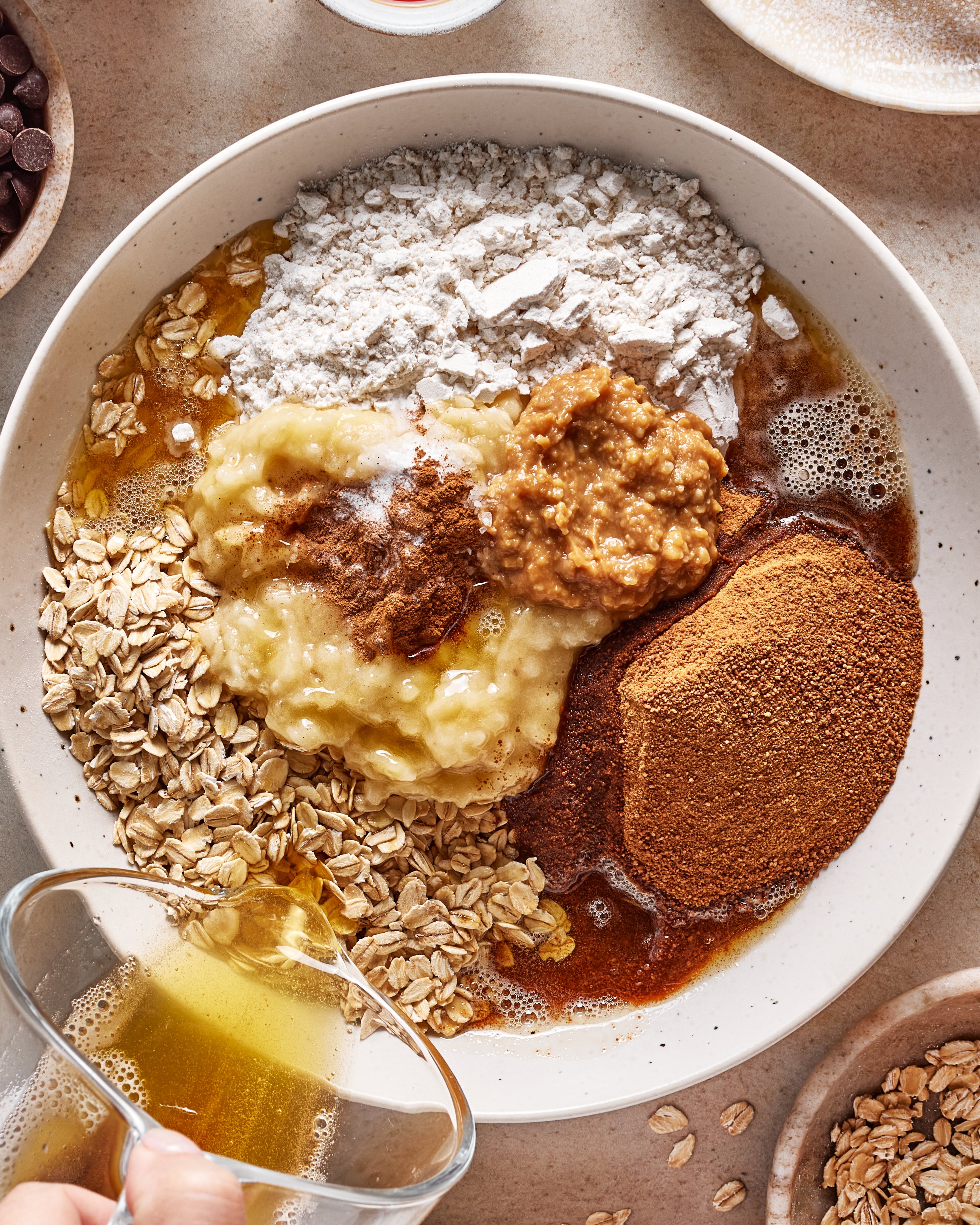A bowl filled with oats, mashed banana, flour, nut butter, coconut sugar, and spices, with a hand pouring oil from a glass cup. Other ingredients are visible in small bowls around the main bowl.