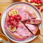A round cake with pink glaze, topped with fresh raspberries and lemon slices, sits on a white plate. A slice is being served, and raspberries, lemons, and plates with forks are nearby on a tiled surface.