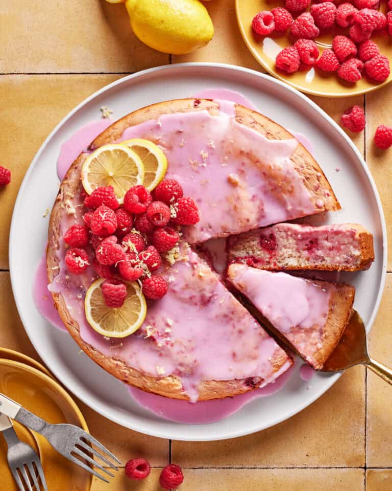 A round cake with pink glaze, topped with fresh raspberries and lemon slices, sits on a white plate. A slice is being served, and raspberries, lemons, and plates with forks are nearby on a tiled surface.