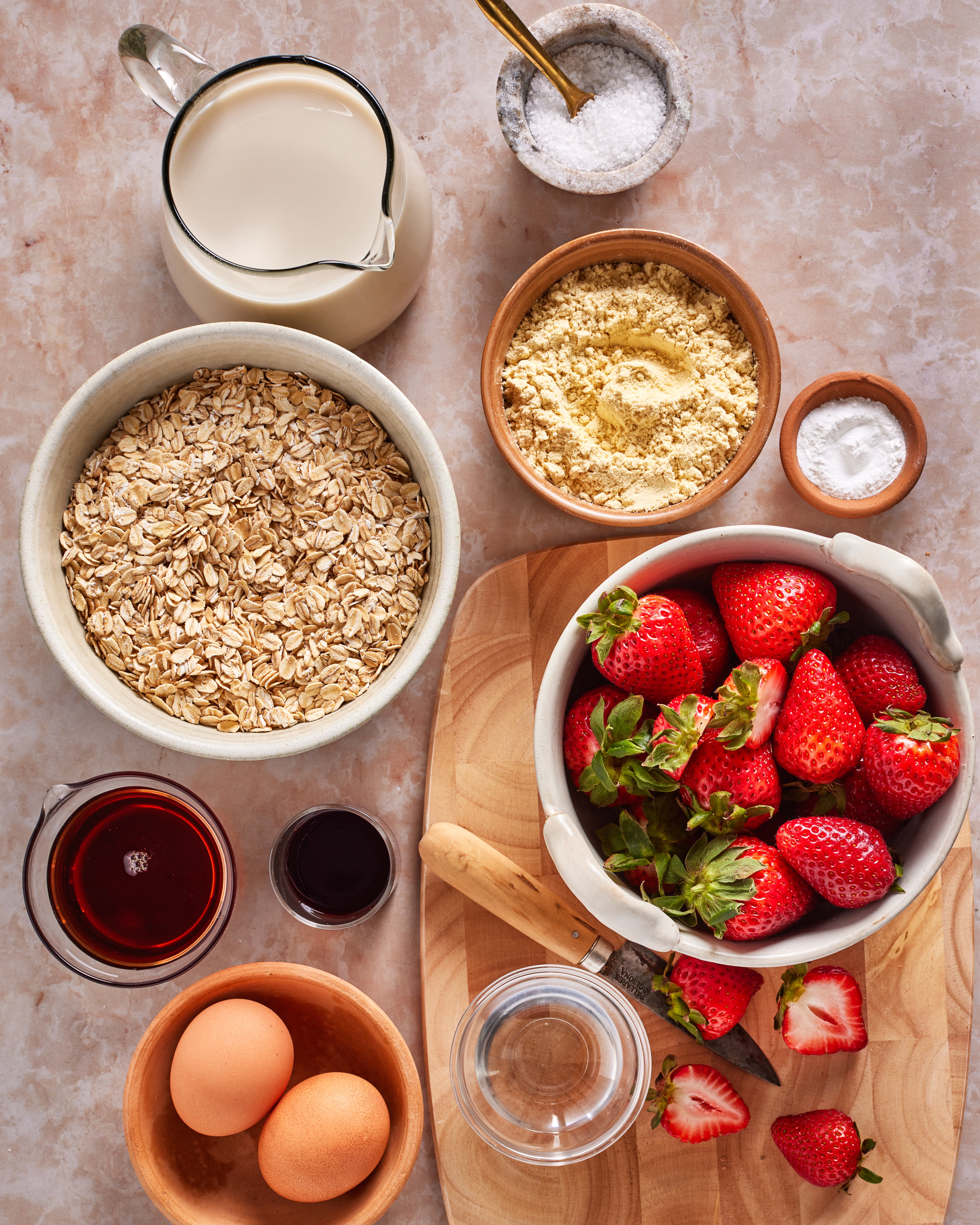 An overhead view of baking ingredients: a bowl of rolled oats, a bowl of strawberries, eggs, milk, maple syrup, vanilla, salt, baking powder, and almond flour on a marble surface with a wooden cutting board.