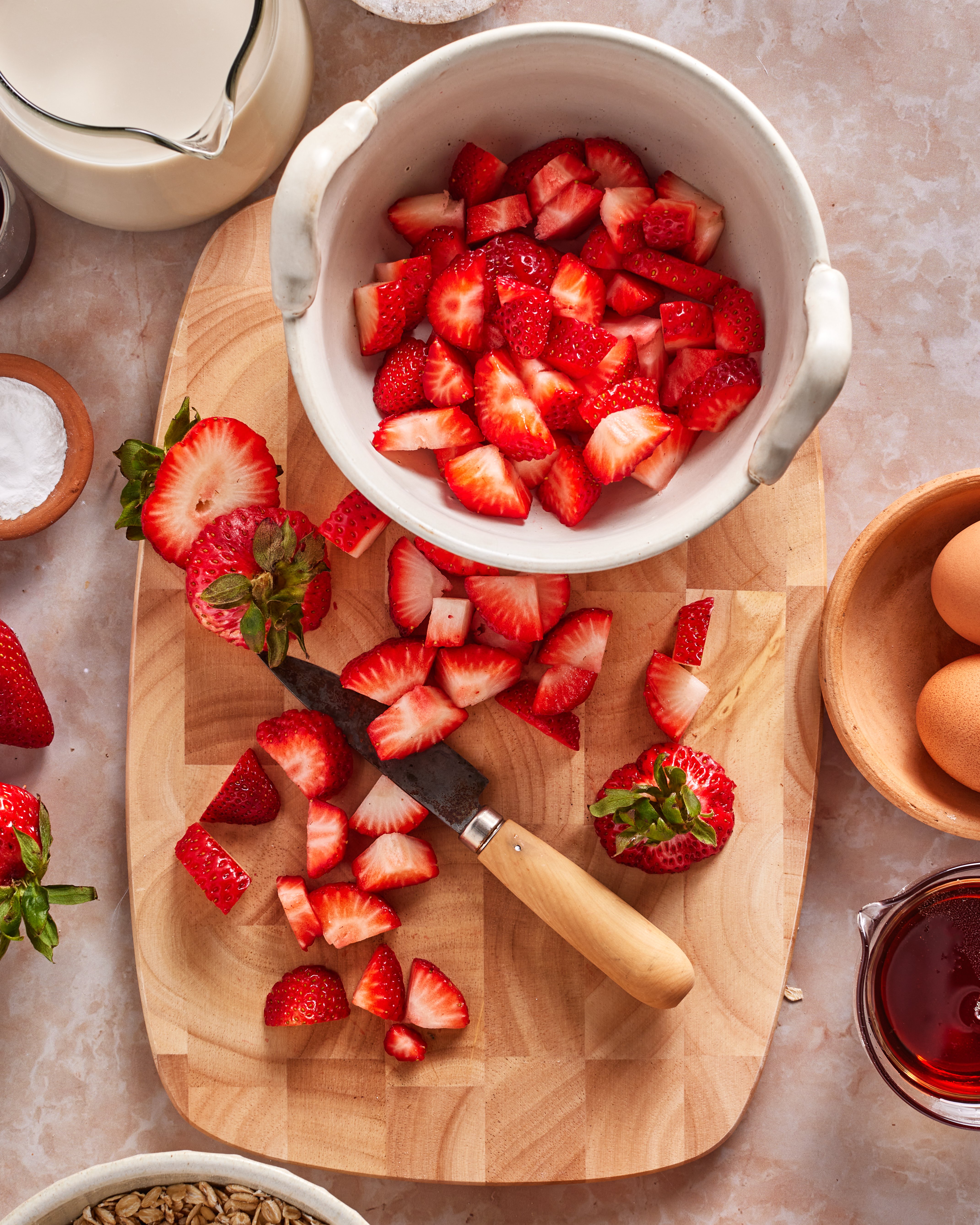 A wooden cutting board with chopped strawberries, a knife, a bowl of more chopped strawberries, whole eggs, a pitcher of milk, a bowl of oats, and a small bowl of syrup on a marble countertop.