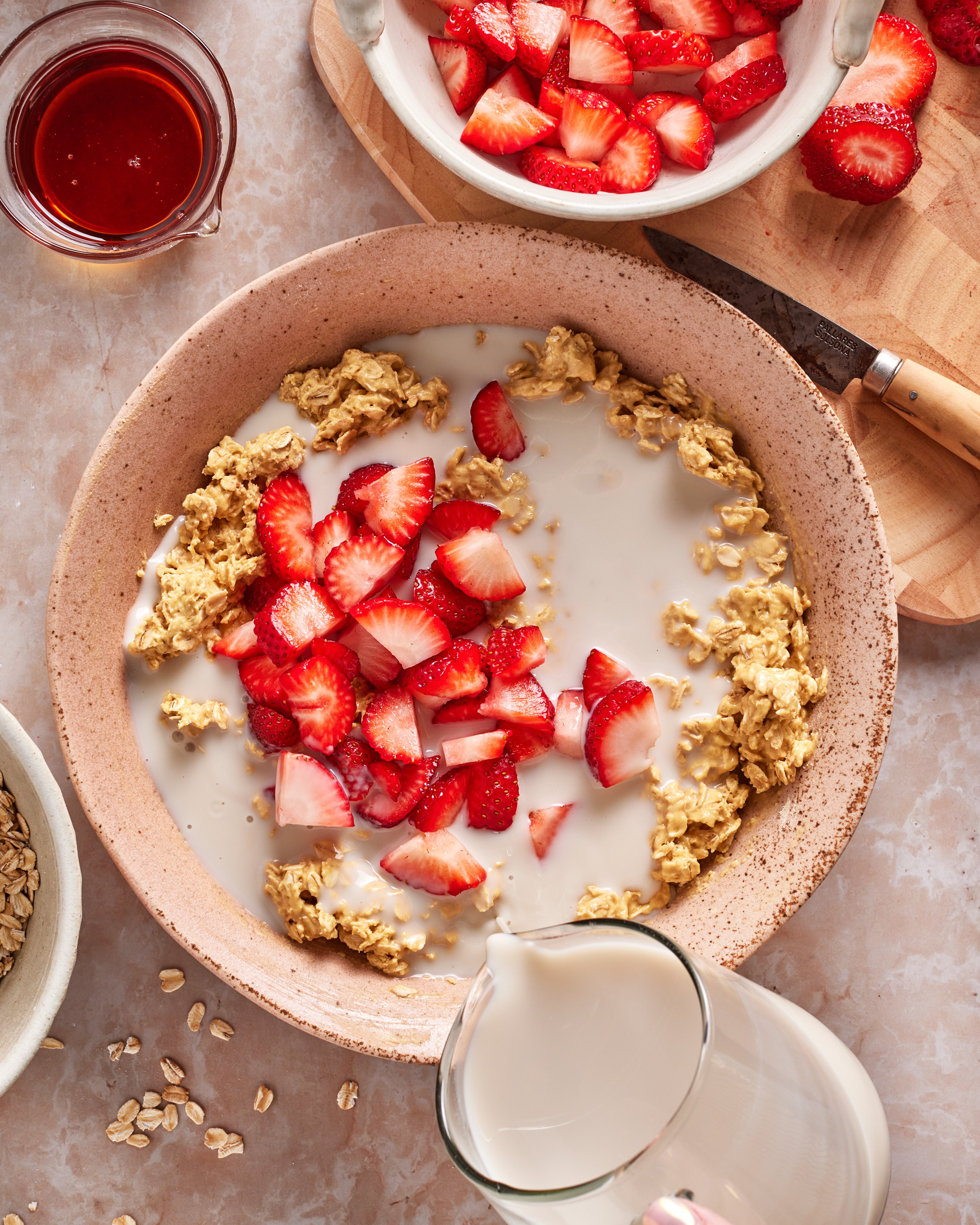 A bowl of granola topped with sliced strawberries and milk being poured in, with extra strawberries, oats, and a small jar of syrup nearby on a light countertop.