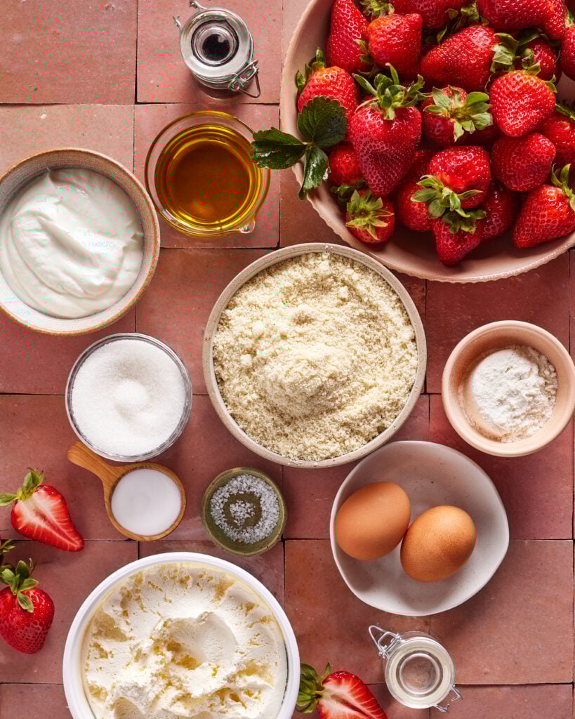 A flat lay of baking ingredients on a tiled surface, including fresh strawberries, eggs, sugar, flour, almond flour, yogurt, oil, baking powder, salt, and a small jar of vanilla extract.