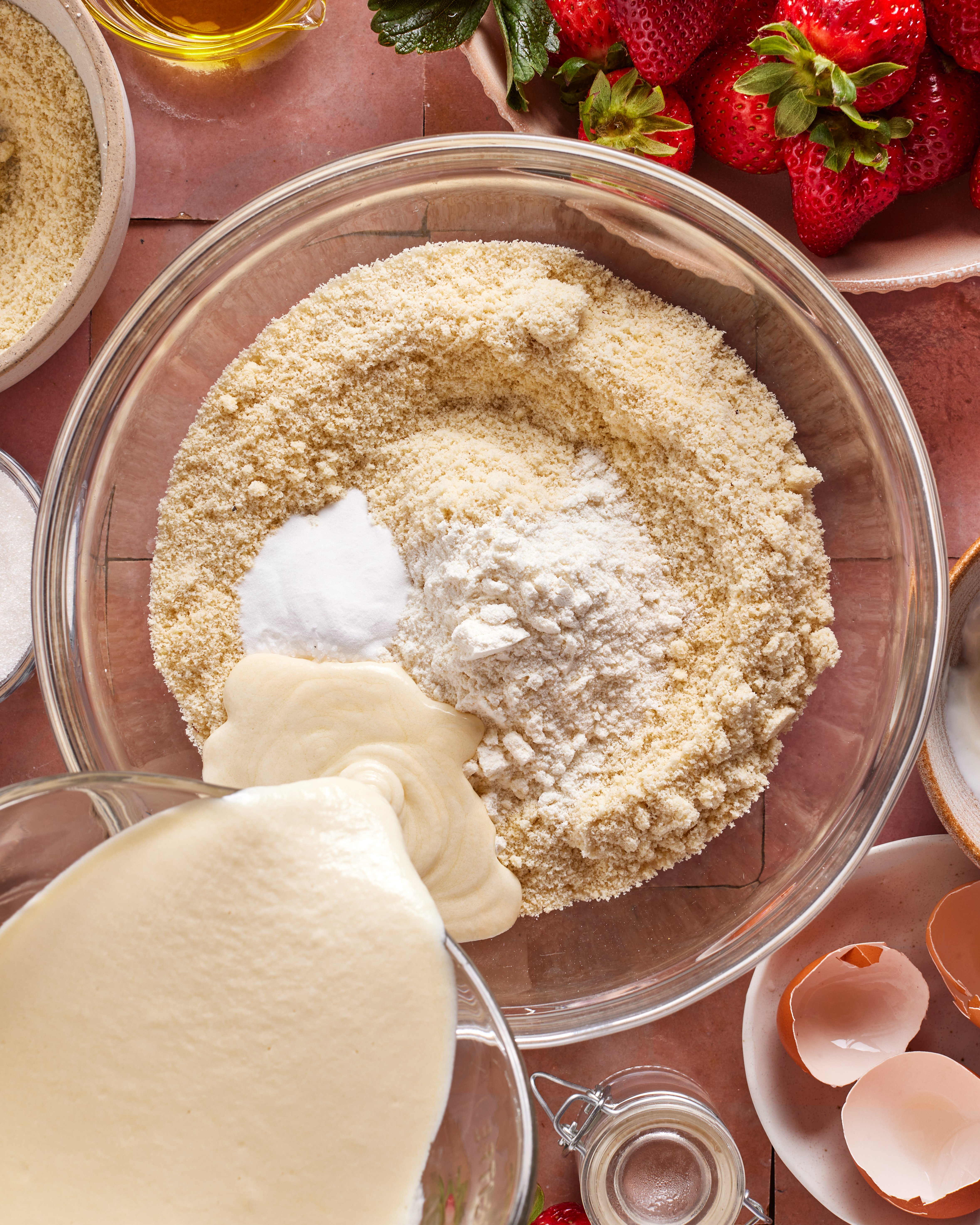 A mixing bowl with almond flour, baking powder, and flour. Batter is being poured in. Surrounding the bowl are strawberries, eggshells, olive oil, and more ingredients on a tiled surface.