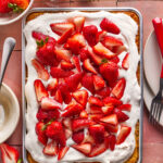 A rectangular sheet cake topped with whipped cream and fresh sliced strawberries sits on a pink tile surface. Plates, forks, a knife, and bowls of strawberries surround the cake.