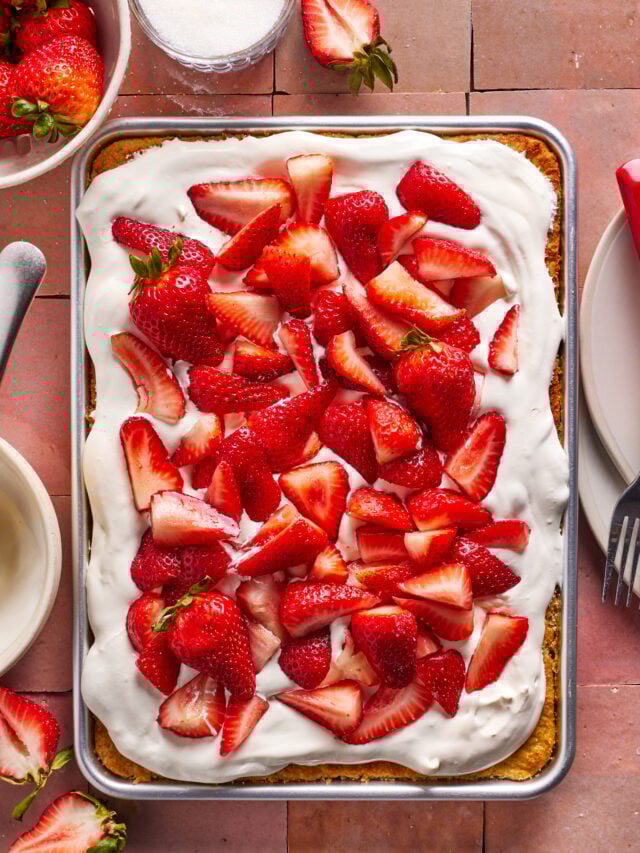 A rectangular sheet cake topped with whipped cream and fresh sliced strawberries sits on a pink tile surface. Plates, forks, a knife, and bowls of strawberries surround the cake.