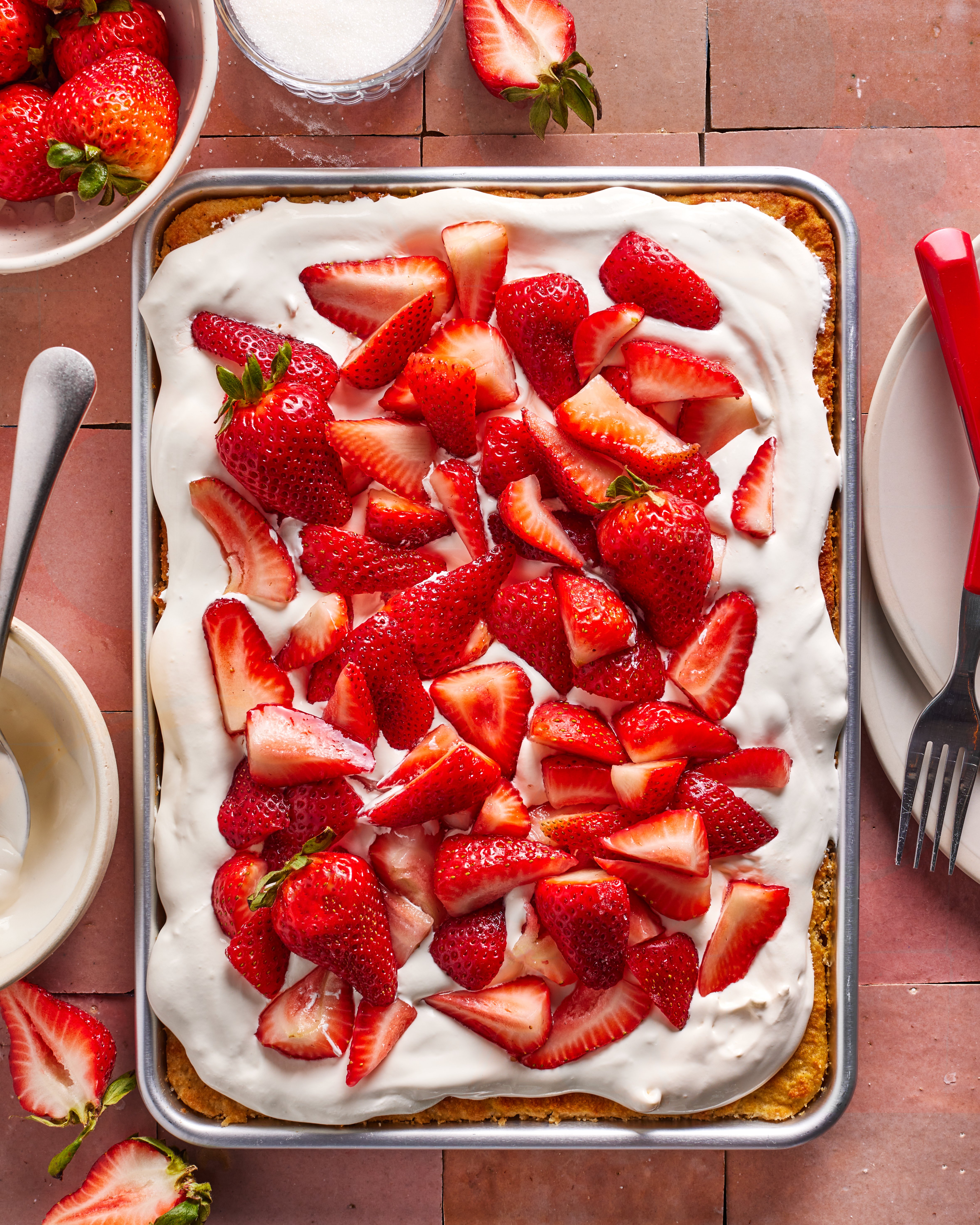 A rectangular sheet cake topped with whipped cream and fresh sliced strawberries sits on a pink tile surface. Plates, forks, a knife, and bowls of strawberries surround the cake.