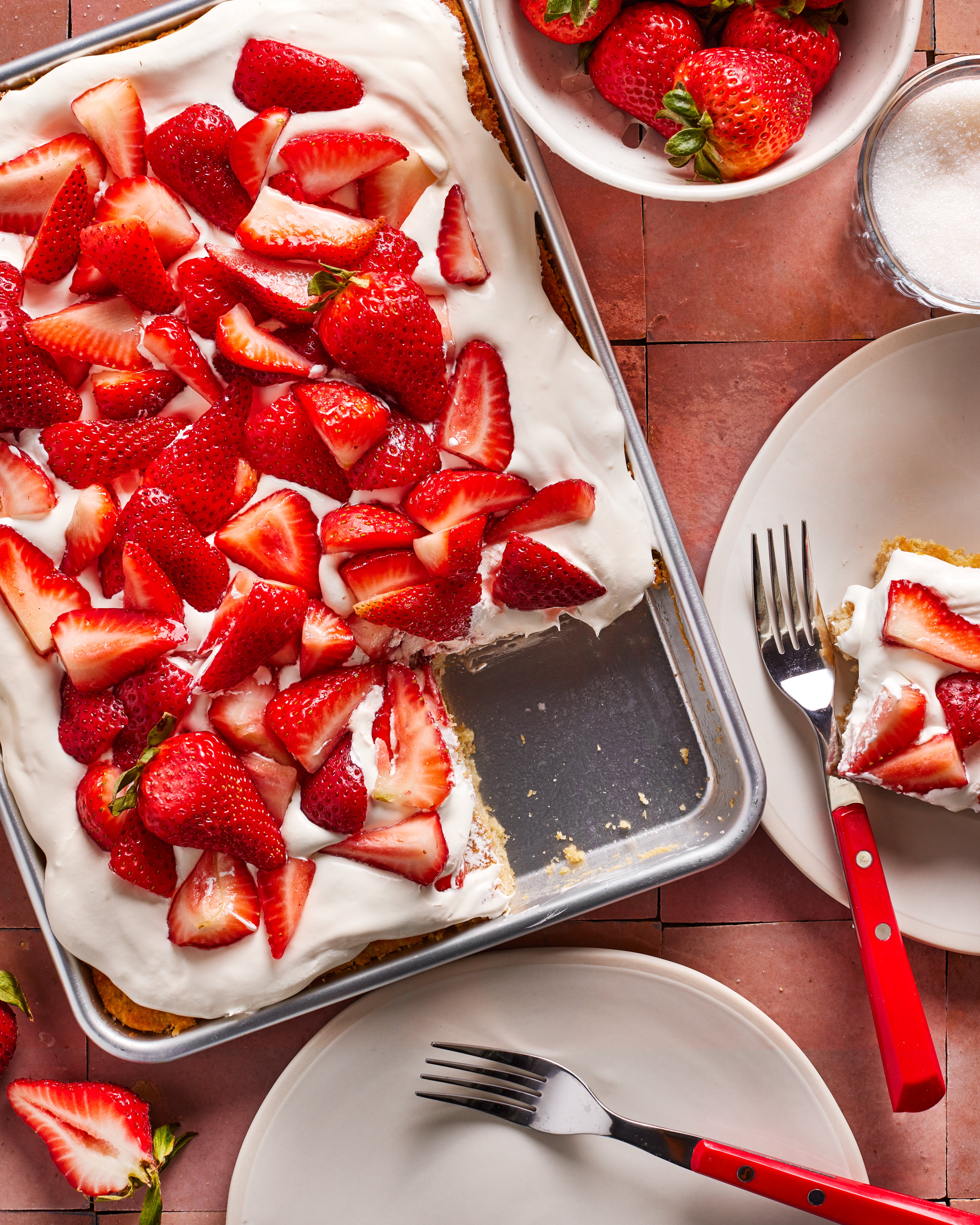 A rectangular cake topped with whipped cream and sliced fresh strawberries sits in a baking pan. Two plates with slices of the cake, forks, and a bowl of strawberries are arranged nearby on a pink tiled surface.