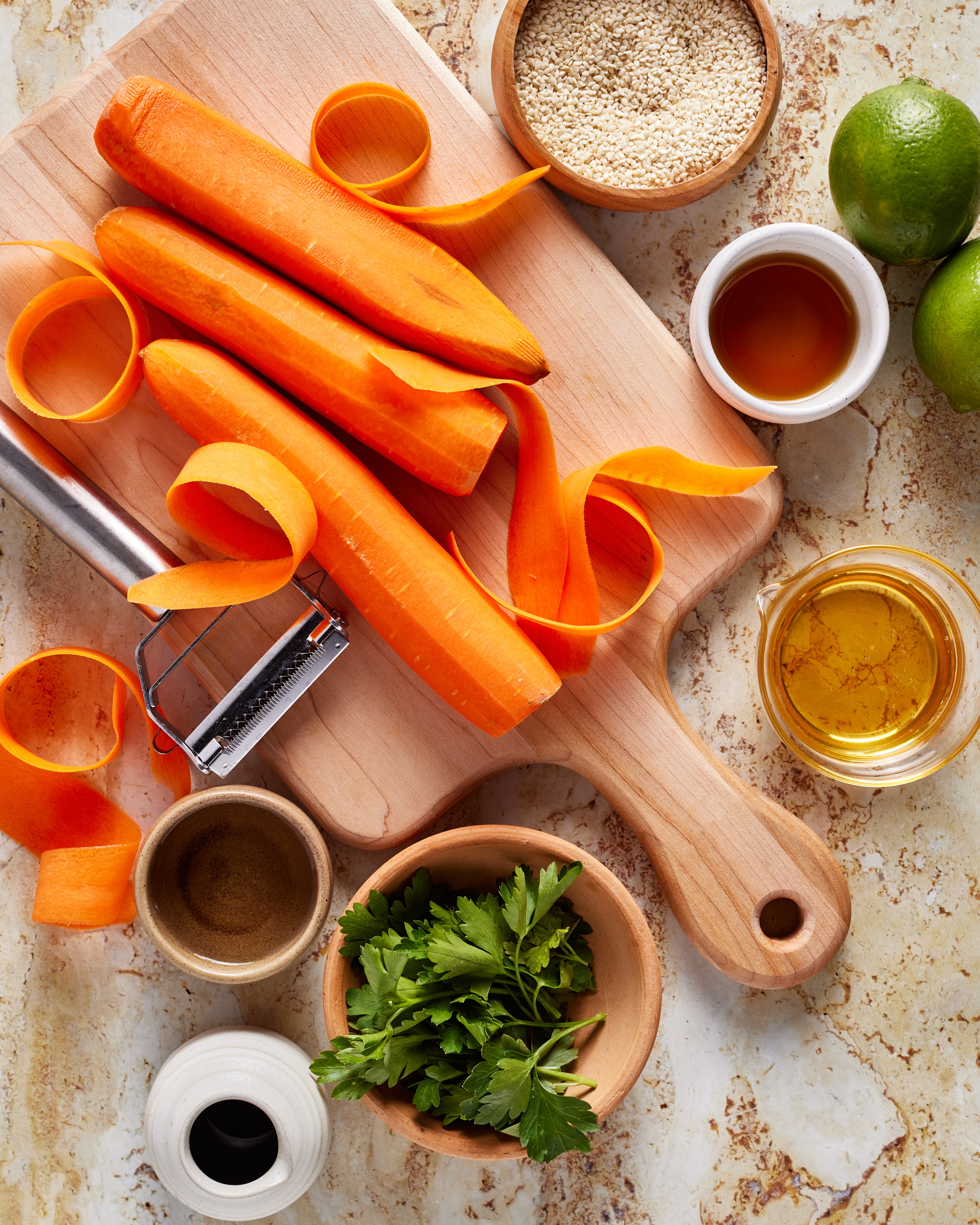 Carrots being peeled on a wooden cutting board, surrounded by a peeler, bowls of sesame seeds, fresh parsley, vinegar, oil, soy sauce, and whole limes on a marble countertop.