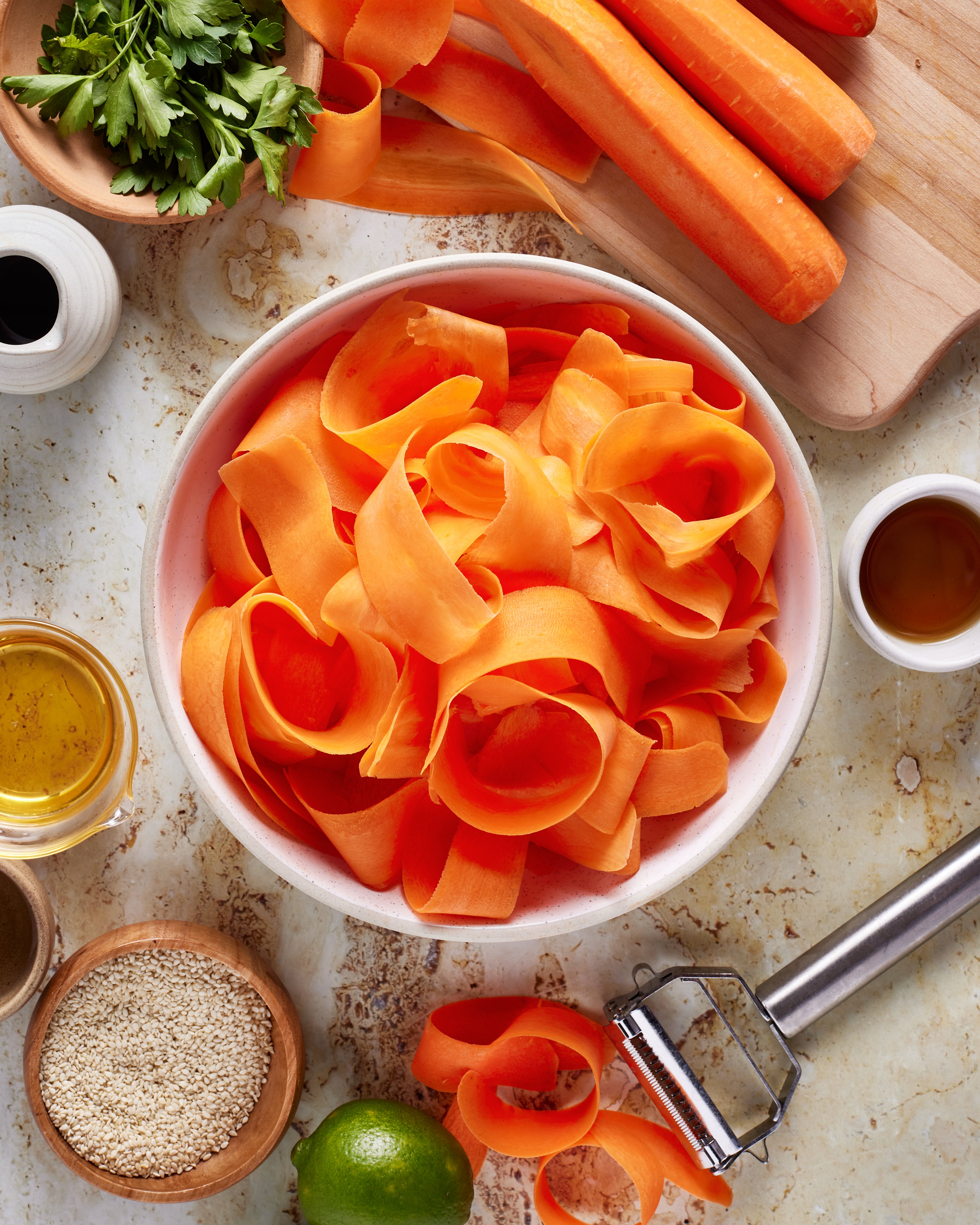 A bowl of wide carrot ribbons surrounded by whole carrots, a vegetable peeler, sesame seeds, fresh parsley, lime, and small bowls of oil and vinegar on a light countertop.