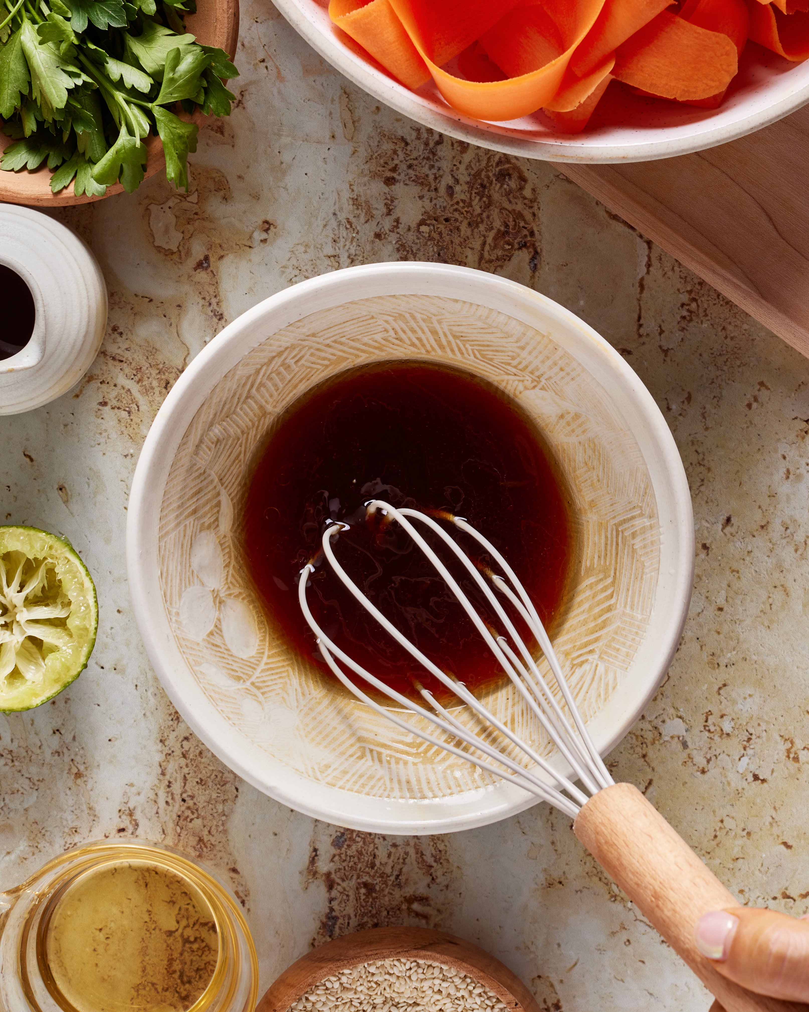 A bowl with dark liquid and a whisk inside, surrounded by a squeezed lime, carrot ribbons, parsley, a small jug, and other ingredients on a marble countertop.