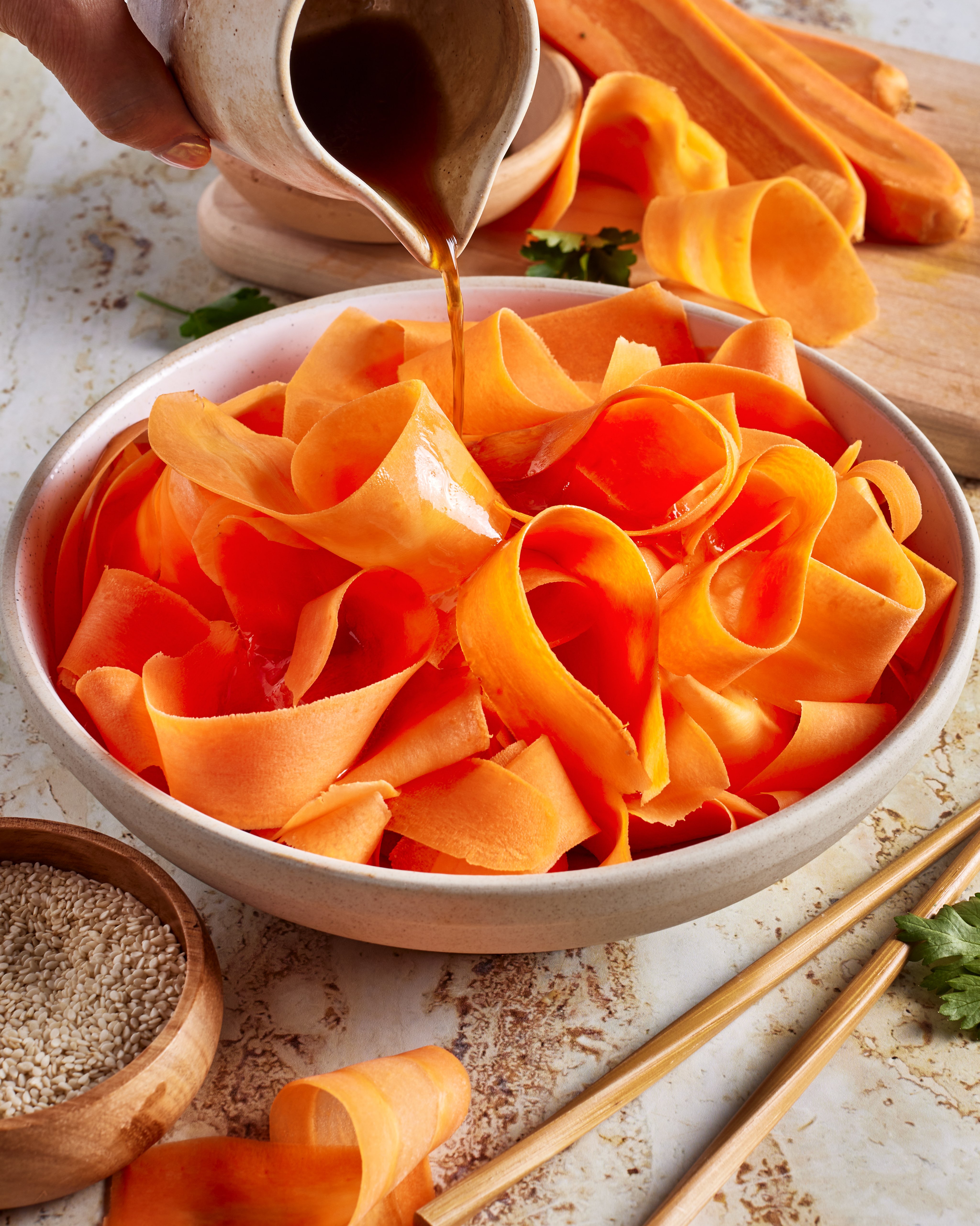 A hand pours brown dressing over wide carrot ribbons in a bowl. Sesame seeds, chopsticks, and fresh carrots are nearby on a textured surface.