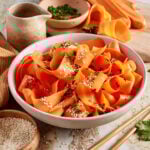 A bowl of carrot ribbons garnished with sesame seeds and chopped herbs sits on a table, surrounded by fresh carrots, herbs, a small pitcher, and a bowl of sesame seeds. Chopsticks lie beside the bowl.