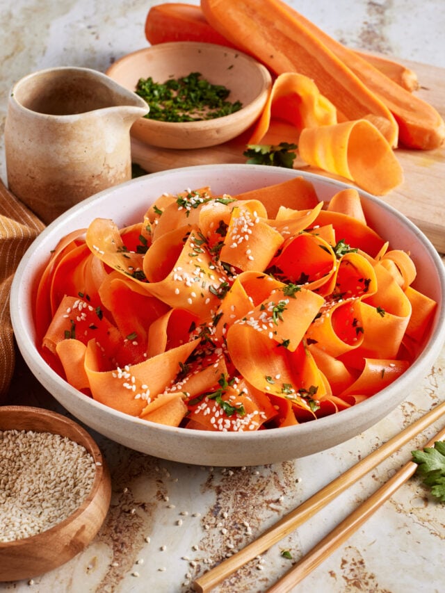 A bowl of carrot ribbons garnished with sesame seeds and chopped herbs sits on a table, surrounded by fresh carrots, herbs, a small pitcher, and a bowl of sesame seeds. Chopsticks lie beside the bowl.