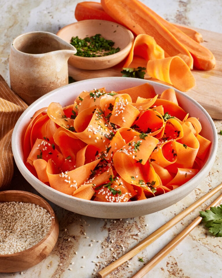 A bowl of carrot ribbons garnished with sesame seeds and chopped herbs sits on a table, surrounded by fresh carrots, herbs, a small pitcher, and a bowl of sesame seeds. Chopsticks lie beside the bowl.