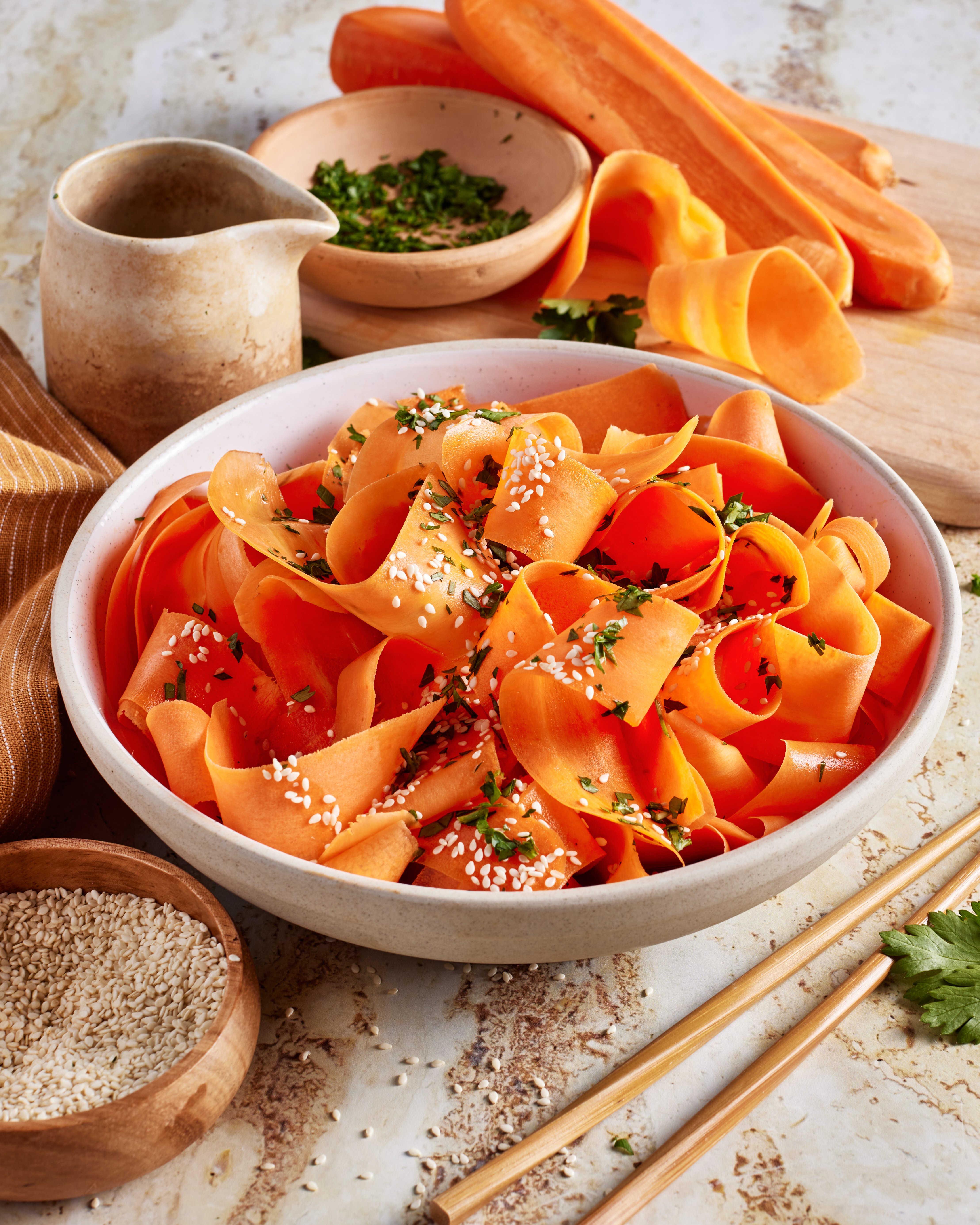 A bowl of carrot ribbons garnished with sesame seeds and chopped herbs sits on a table, surrounded by fresh carrots, herbs, a small pitcher, and a bowl of sesame seeds. Chopsticks lie beside the bowl.
