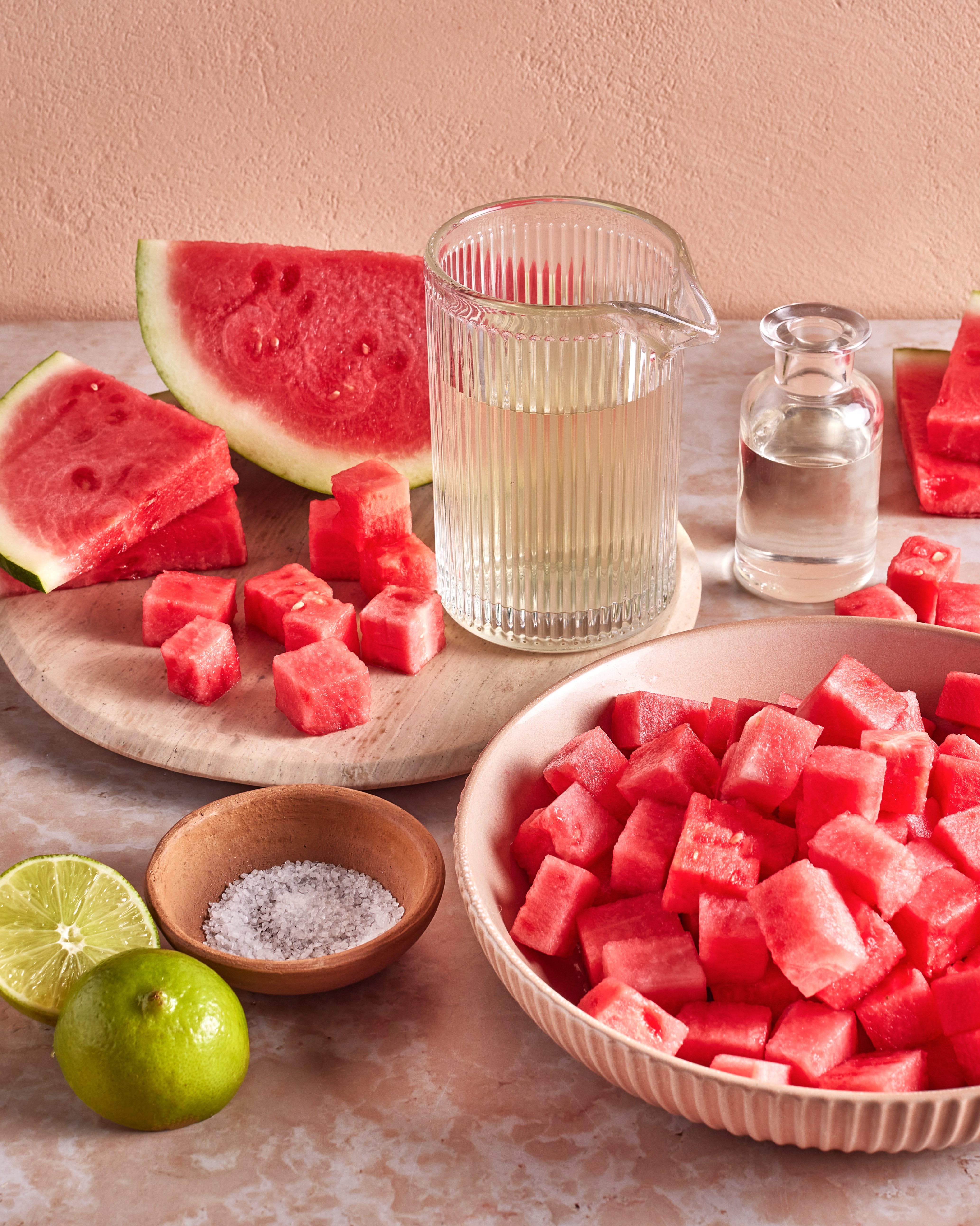 Cubes and slices of watermelon on plates, a glass pitcher of light juice, a small bottle of clear liquid, lime halves, and a small bowl of salt on a light stone surface with a peach-colored background.