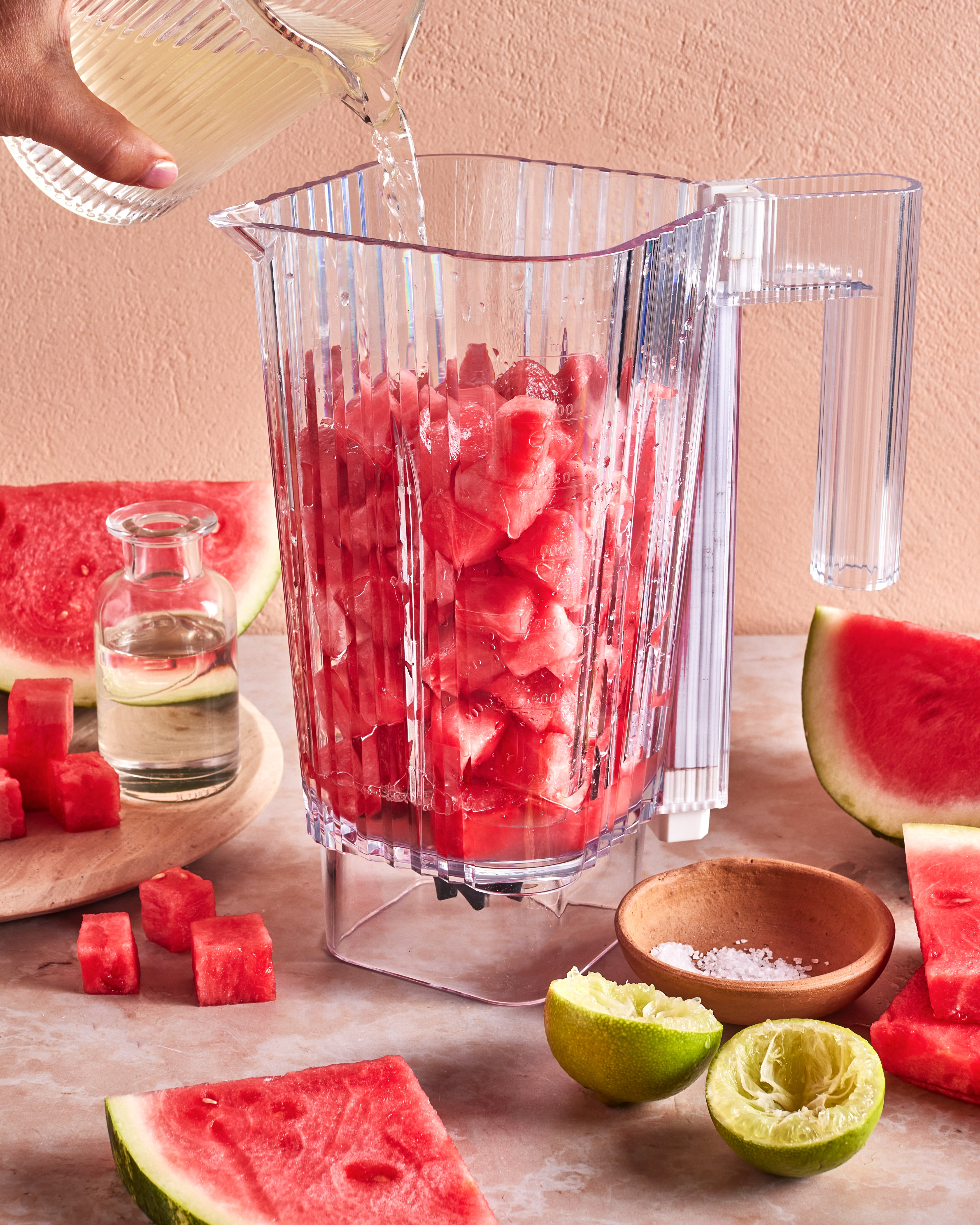 A blender pitcher filled with watermelon chunks is being topped with liquid. Surrounding the pitcher are cut watermelon, a halved lime, a bowl of salt, and a small bottle of clear liquid on a countertop.
