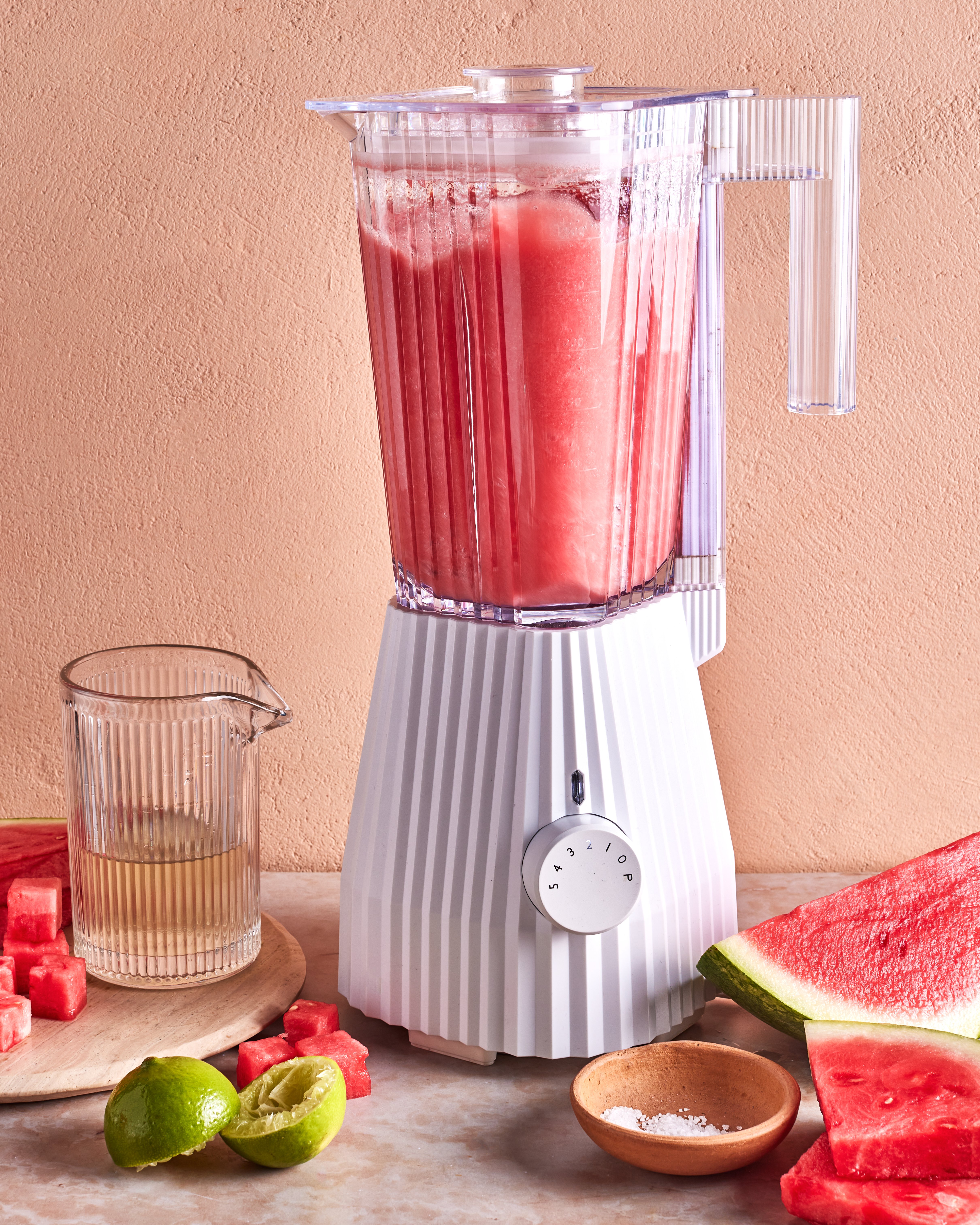 A white blender filled with pink watermelon juice sits on a counter, surrounded by a glass pitcher of juice, a cut lime, a bowl of salt, and fresh watermelon slices.