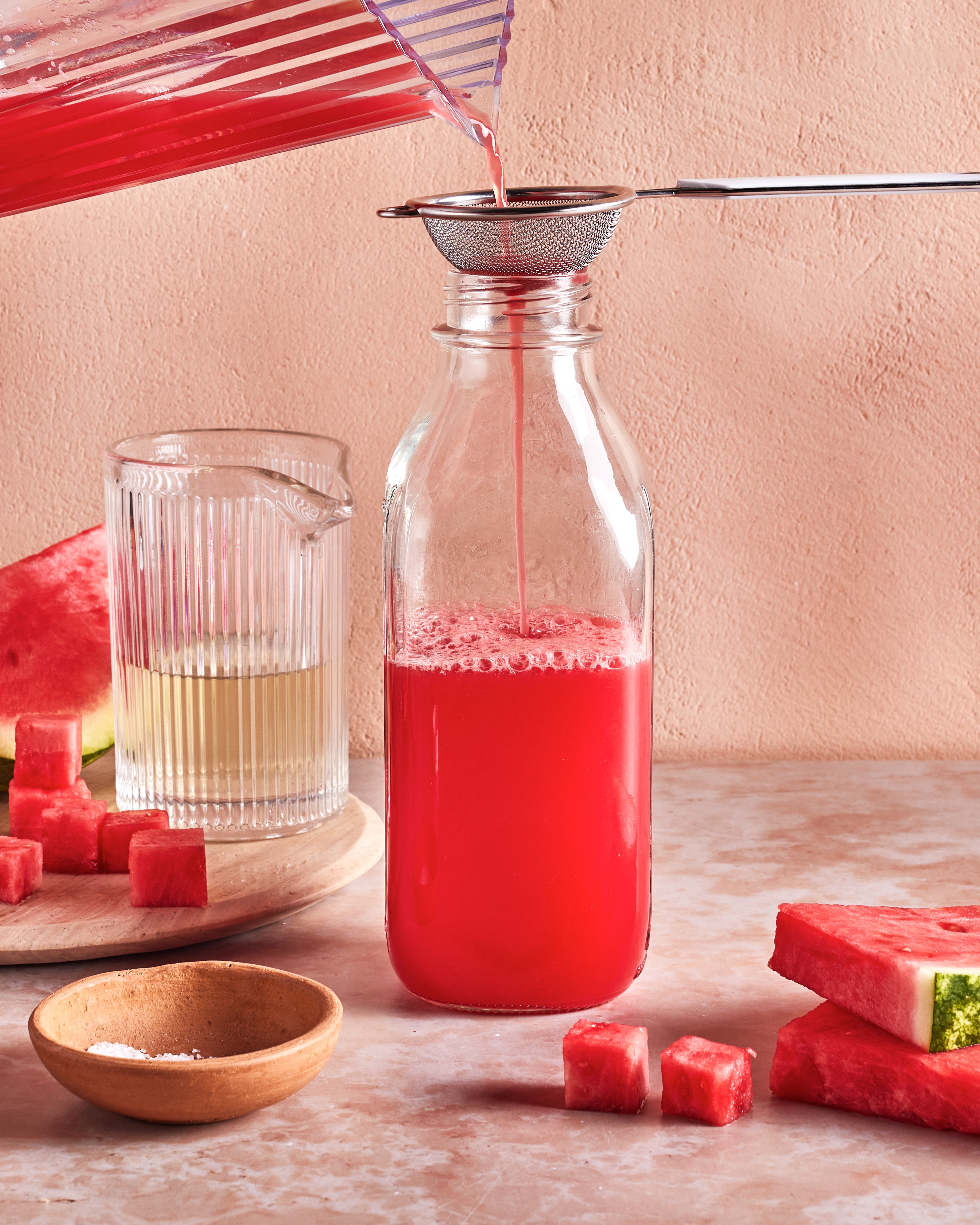 A jug pours bright red watermelon juice through a strainer into a glass bottle. Nearby are a glass of juice, watermelon slices, cubes, and a small bowl of salt, all set on a light pink surface and background.