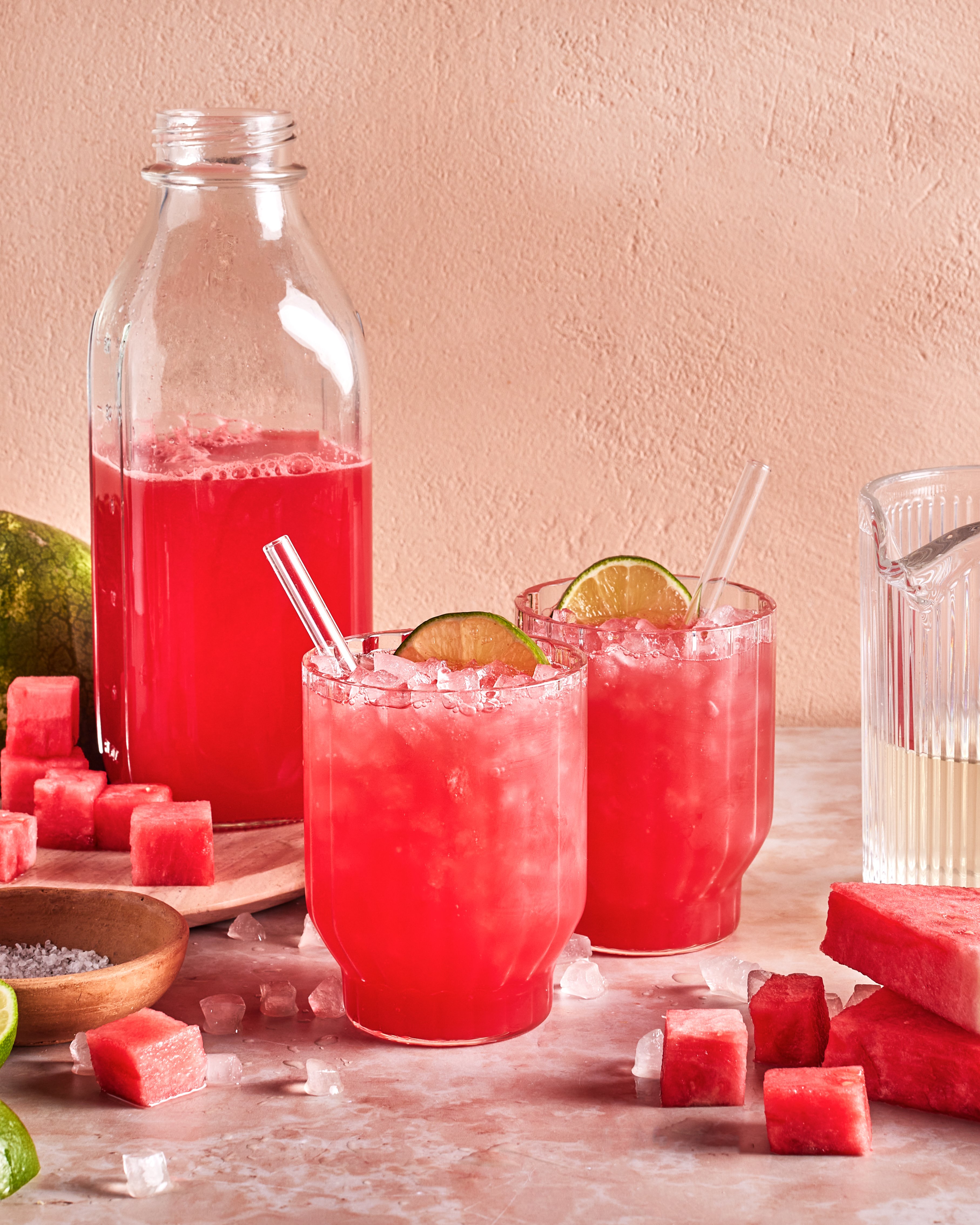 Two glasses filled with a bright pink watermelon drink, crushed ice, lime slices, and straws sit on a table with a bottle of the same drink, watermelon cubes, lime, and a pitcher nearby.