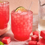 A glass of pink watermelon cocktail with crushed ice, garnished with a lime wheel and a clear straw, sits on a table surrounded by watermelon cubes, a lime, and a glass pitcher.