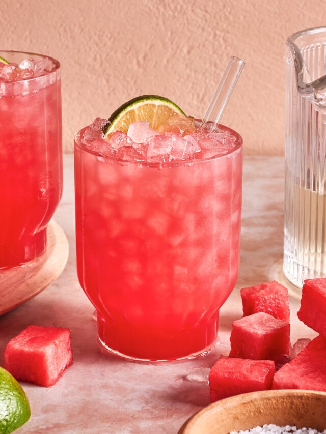 A glass of pink watermelon cocktail with crushed ice, garnished with a lime wheel and a clear straw, sits on a table surrounded by watermelon cubes, a lime, and a glass pitcher.