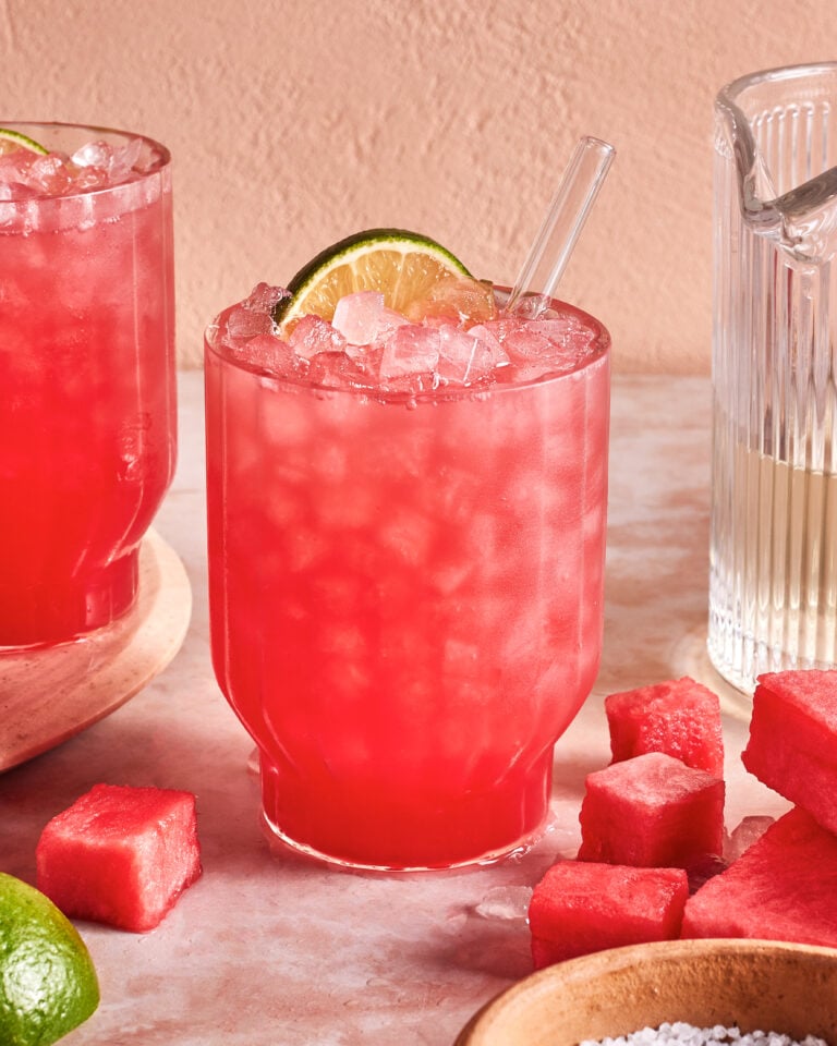 A glass of pink watermelon cocktail with crushed ice, garnished with a lime wheel and a clear straw, sits on a table surrounded by watermelon cubes, a lime, and a glass pitcher.